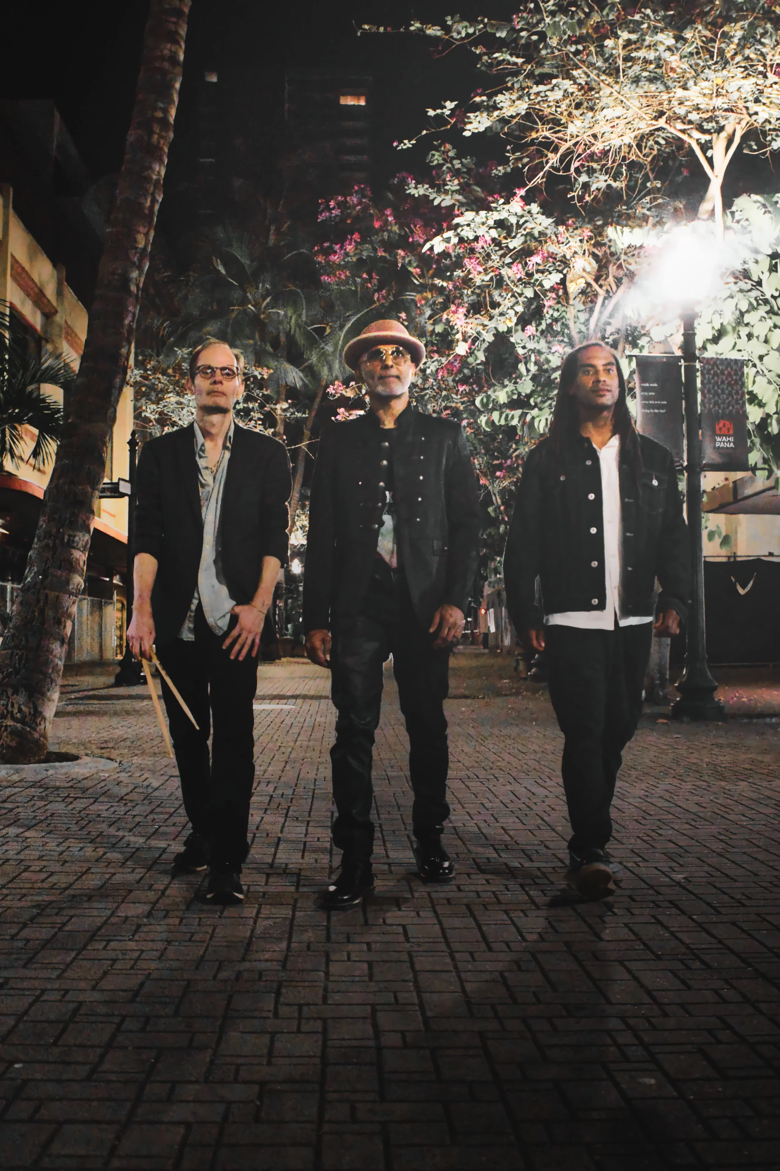 Three men walking on a city street at night, illuminated by streetlights. The man in the center wears a fedora and sunglasses, while the other two men wear casual jackets and jeans.