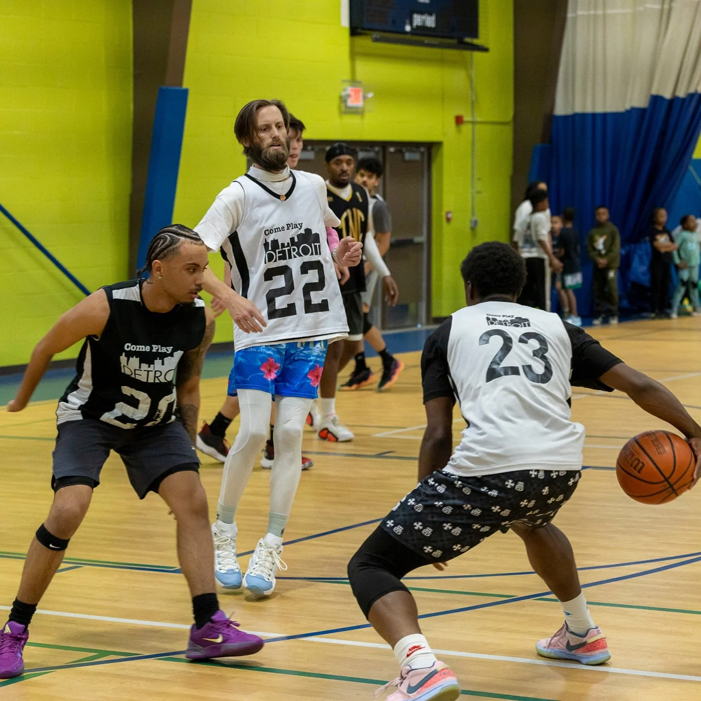 Group of young men playing basketball on an indoor court, with one player dribbling and others defending, while spectators watch in the background.