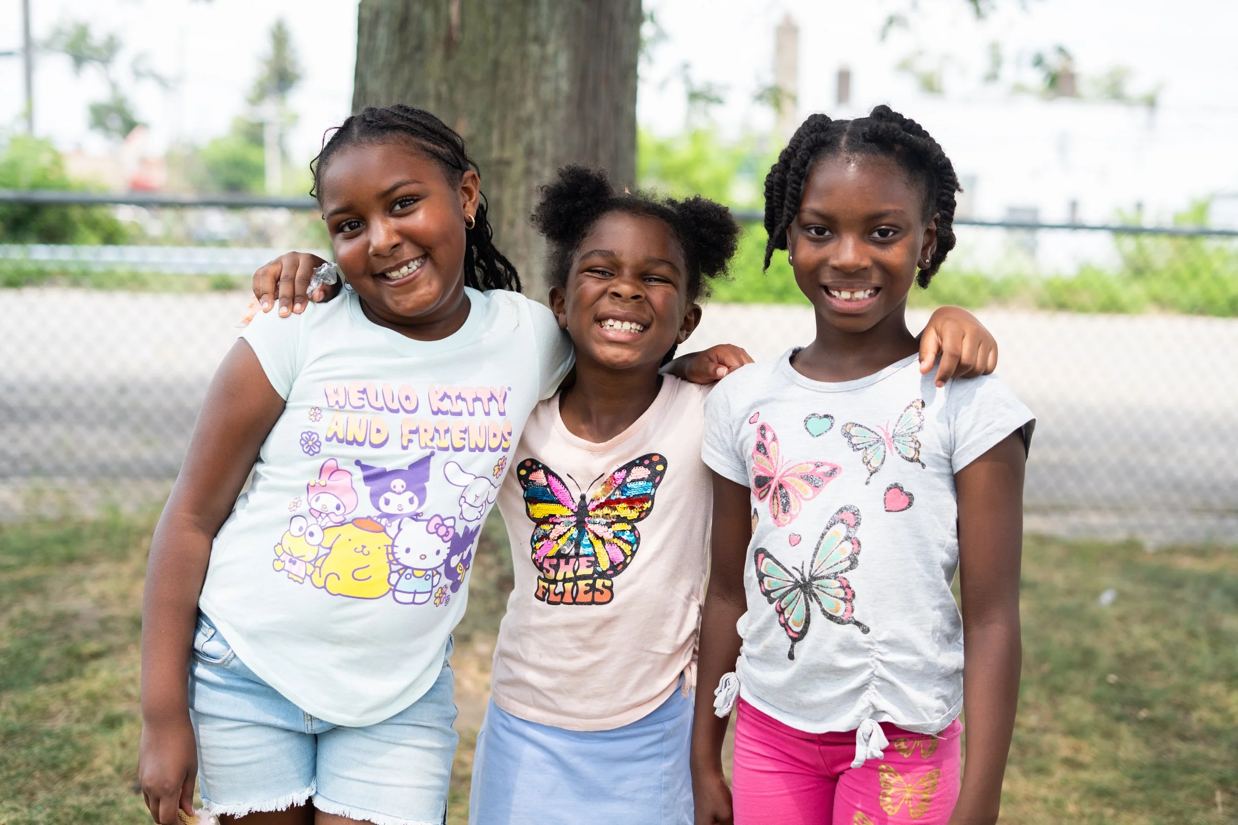 Three young girls standing outside, smiling, with their arms around each other. They are in front of a large tree and a chain-link fence, with greenery in the background.