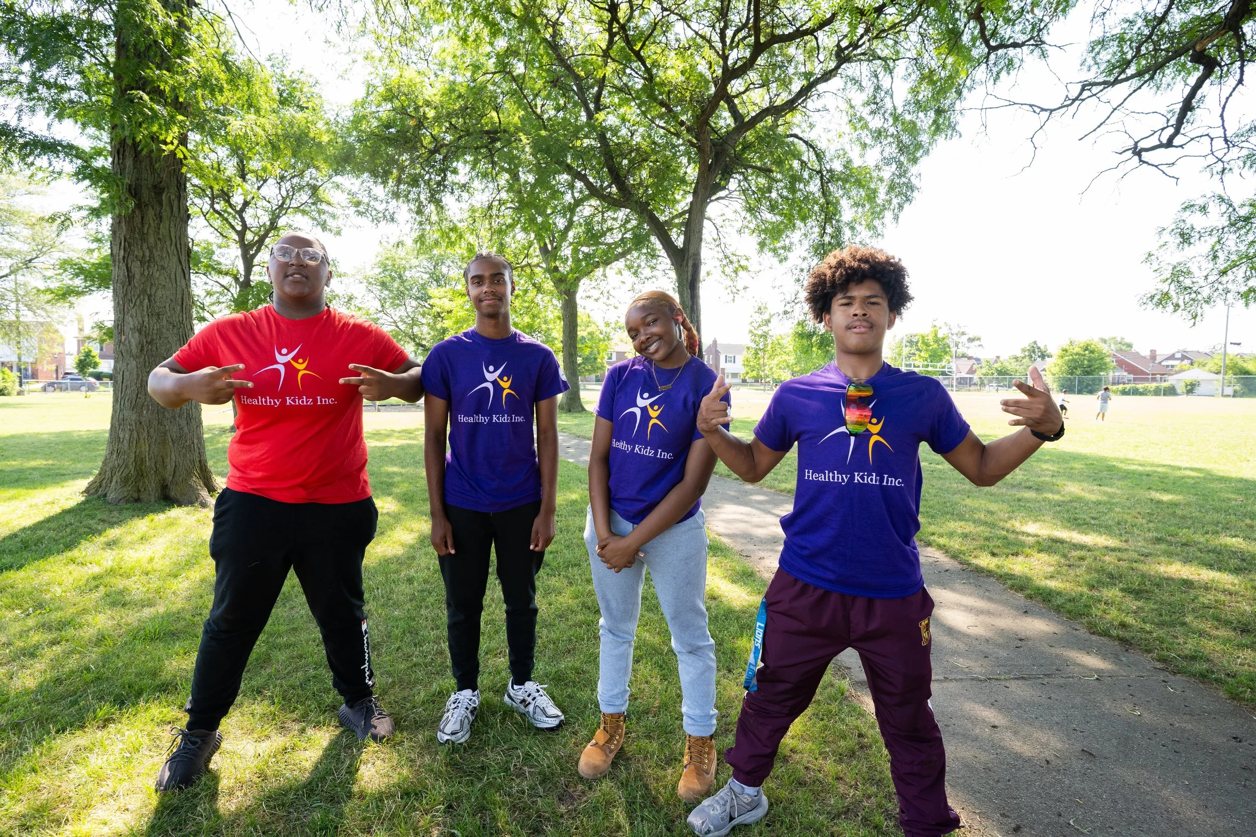 Group of four diverse kids standing outdoors in a park, wearing matching t-shirts with the logo and text "Healthy Kidz Inc." They are posing confidently on a sunny day with green trees and a grassy field in the background.