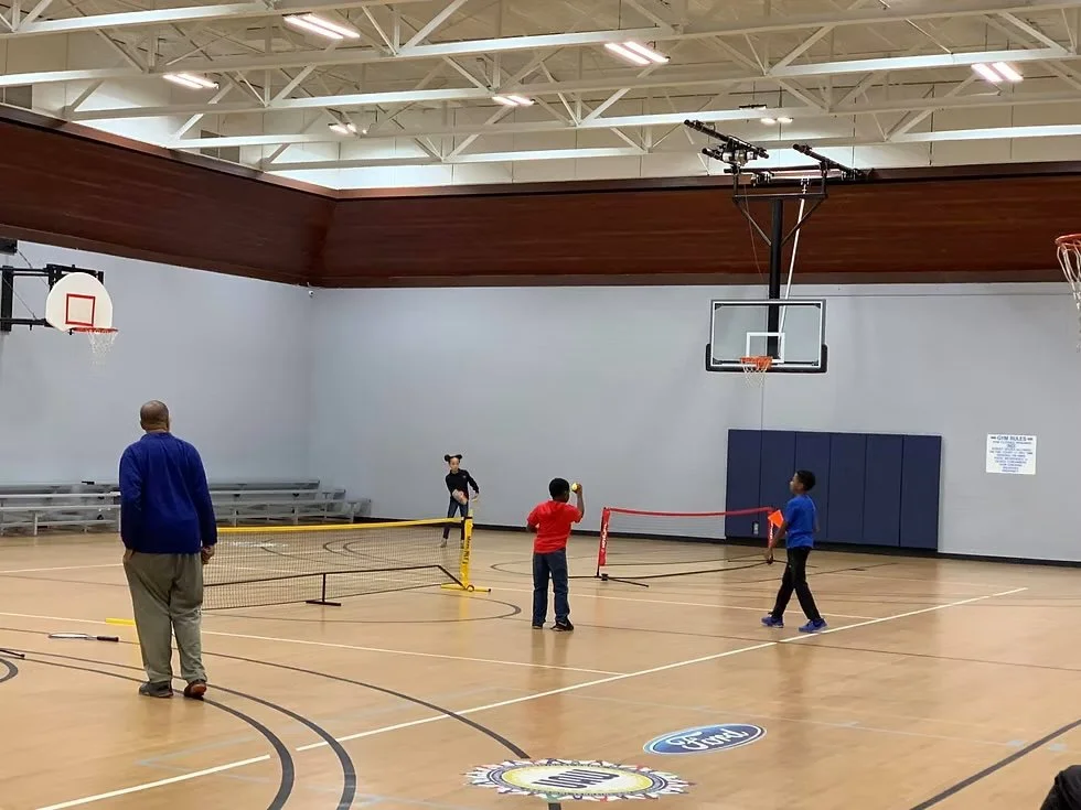 Children playing pickleball in an indoor gymnasium. There are two pickleball nets set up, and a man is observing while three kids are playing and one kid is sitting on the bench.