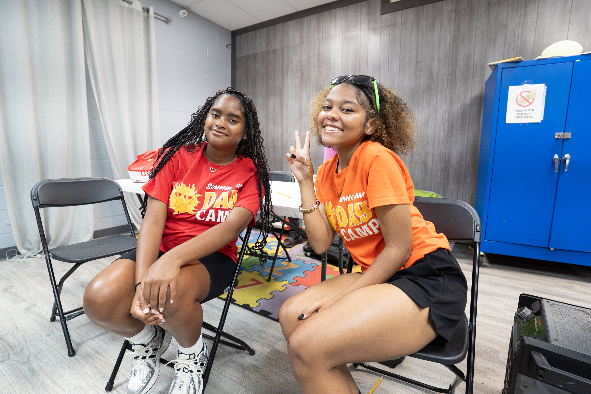 Two girls sitting on black folding chairs indoors, one wearing a red T-shirt and the other an orange T-shirt, both smiling at the camera, one girl making a peace sign. Behind them is a colorful foam puzzle mat, a blue storage cabinet, and a gray wooden wall.