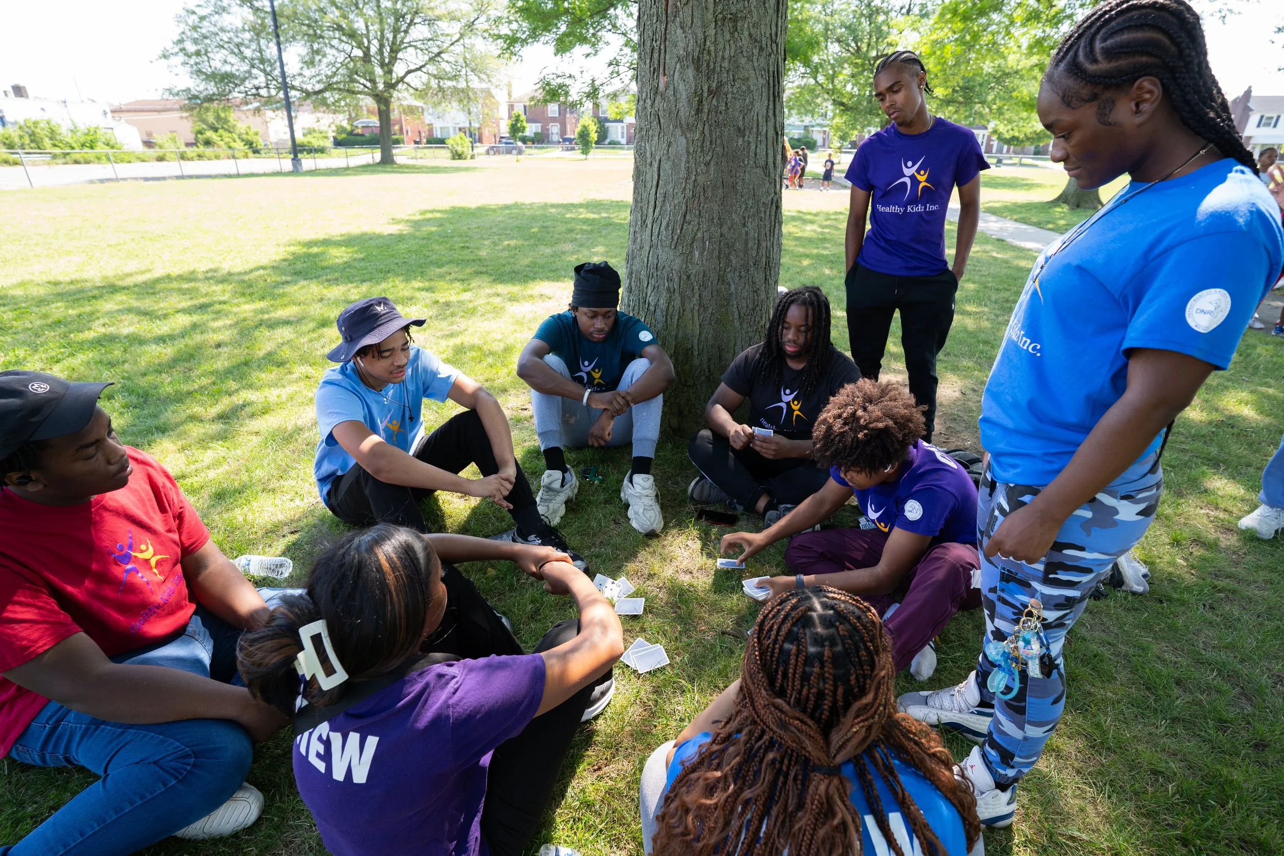 Group of young people sitting and standing in a circle under a tree in a park, engaging in a group activity.