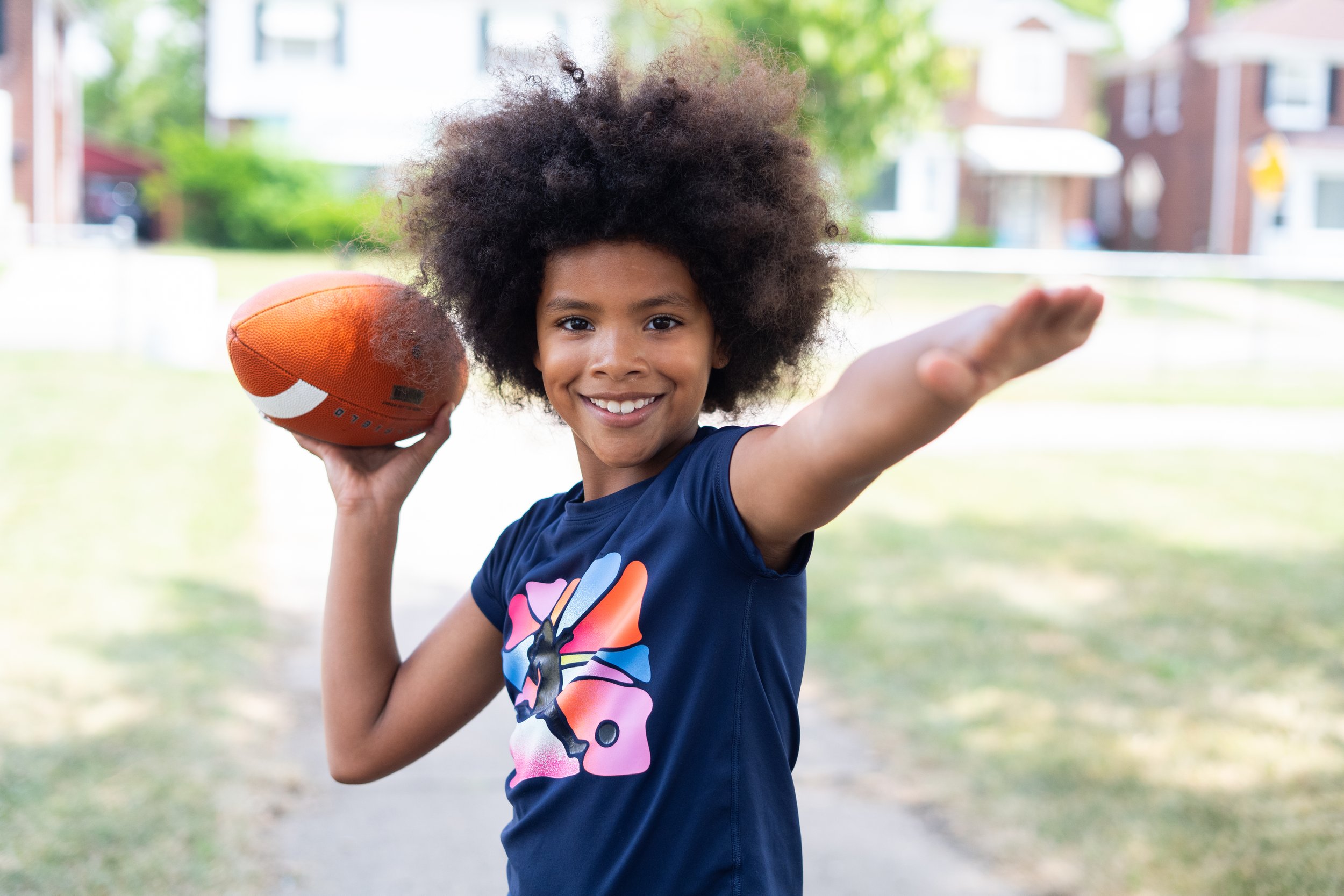 A young girl with curly hair smiling, holding a football in her right hand, and extending her left arm forward.