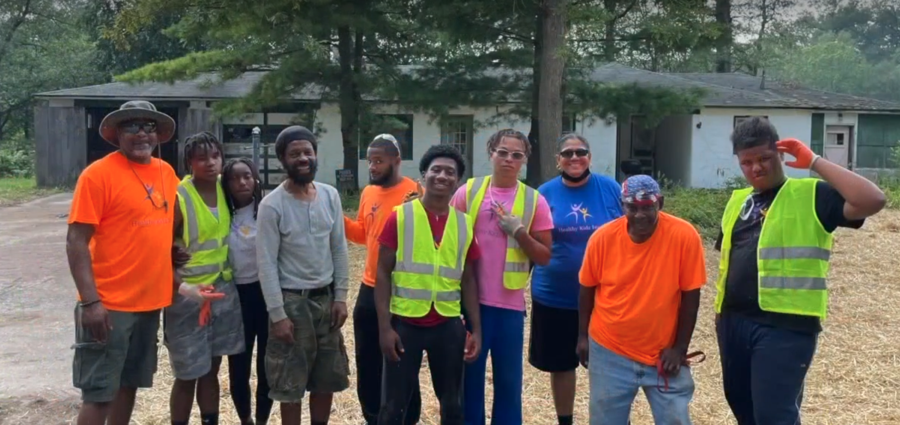 Group of people standing outdoors in front of a building, some wearing safety vests and casual clothing, with trees in the background.