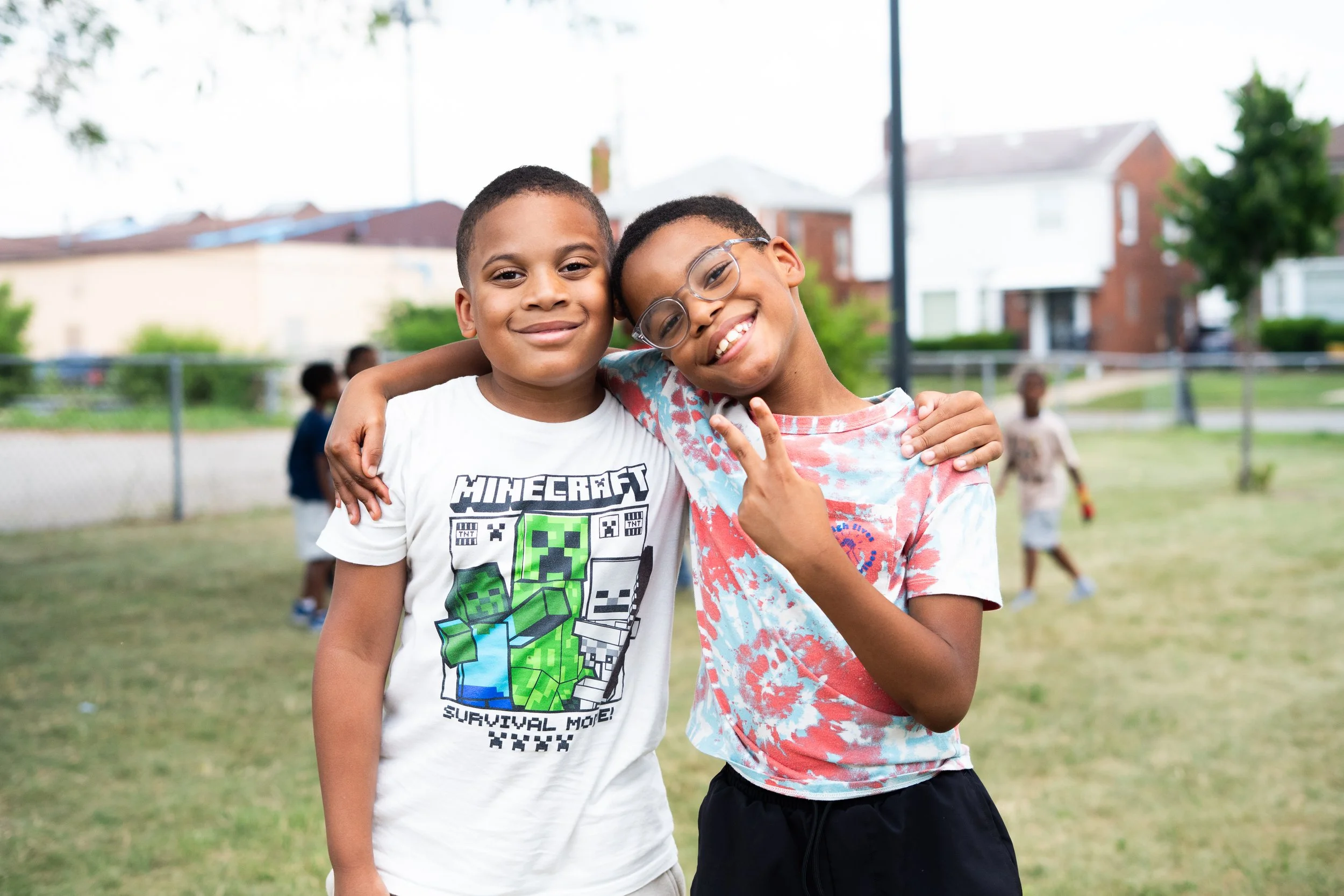 Two boys smiling, one wearing a white Minecraft shirt and the other with glasses and a tie-dye shirt, standing arm in arm outside on a grassy field with a chain-link fence and houses in the background.