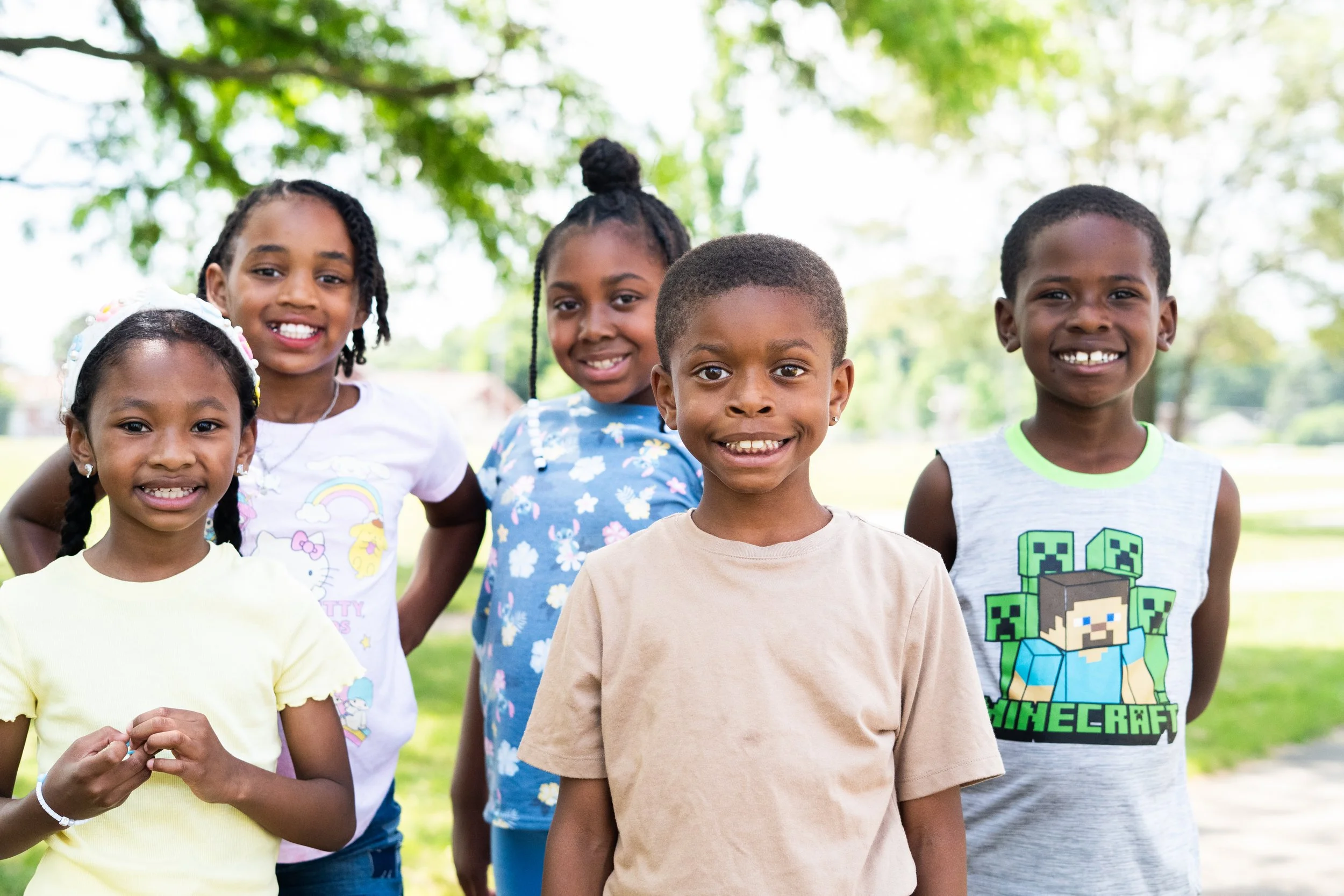 Five smiling children standing outdoors in a park with trees in the background.