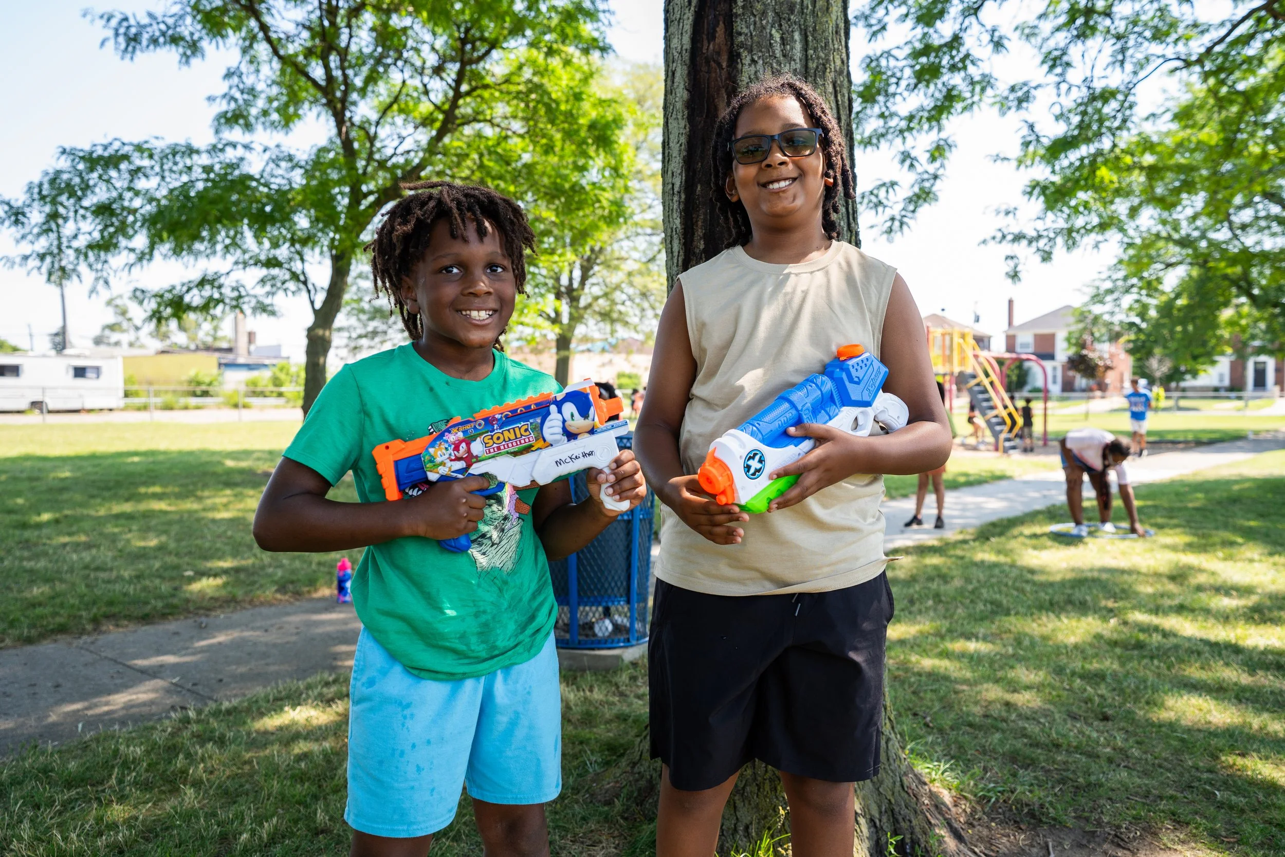 Two children, a boy and a girl, standing outdoors beside a tree, holding toy water guns, smiling, with a park playground and other children playing in the background.