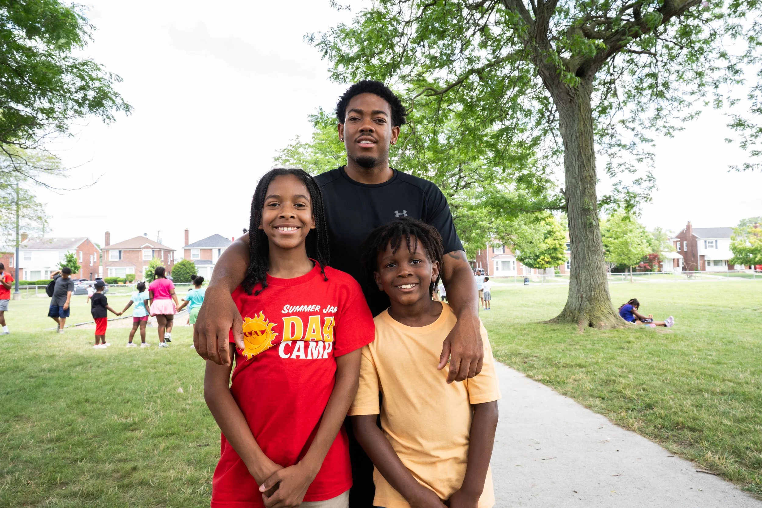 A man and two children smiling outdoors in a park, with other children playing in the background and trees and houses in the distance.