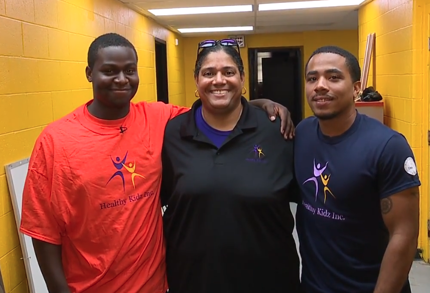 Three smiling individuals standing together in a yellow hallway, wearing shirts with 'Healthy Kidz Inc.' logo.