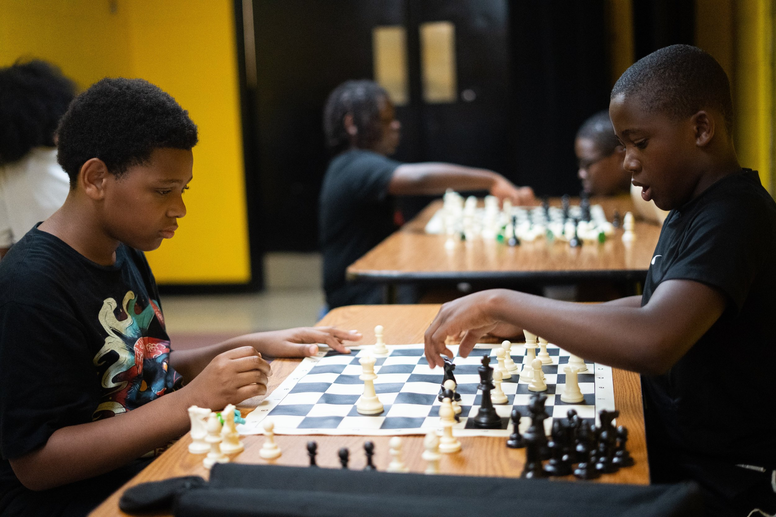 Two boys playing chess at a wooden table while other players play in the background.