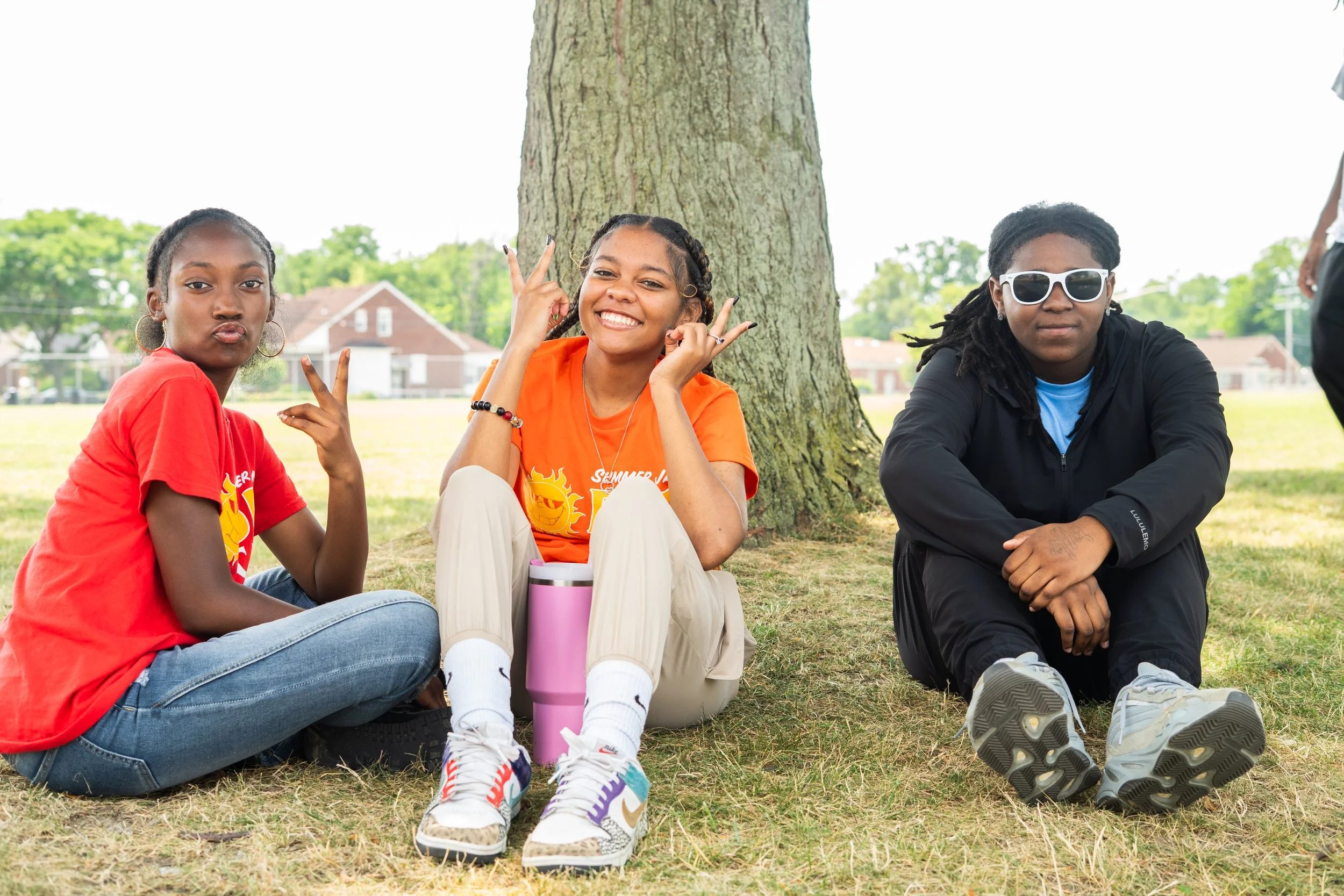 Three young people sitting on the grass near a tree in a park, smiling and making peace and rock hand gestures.