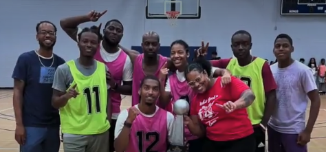 Group of eight young men in basketball jerseys and casual clothes posing in a gymnasium, with basketball hoops and a court in the background.