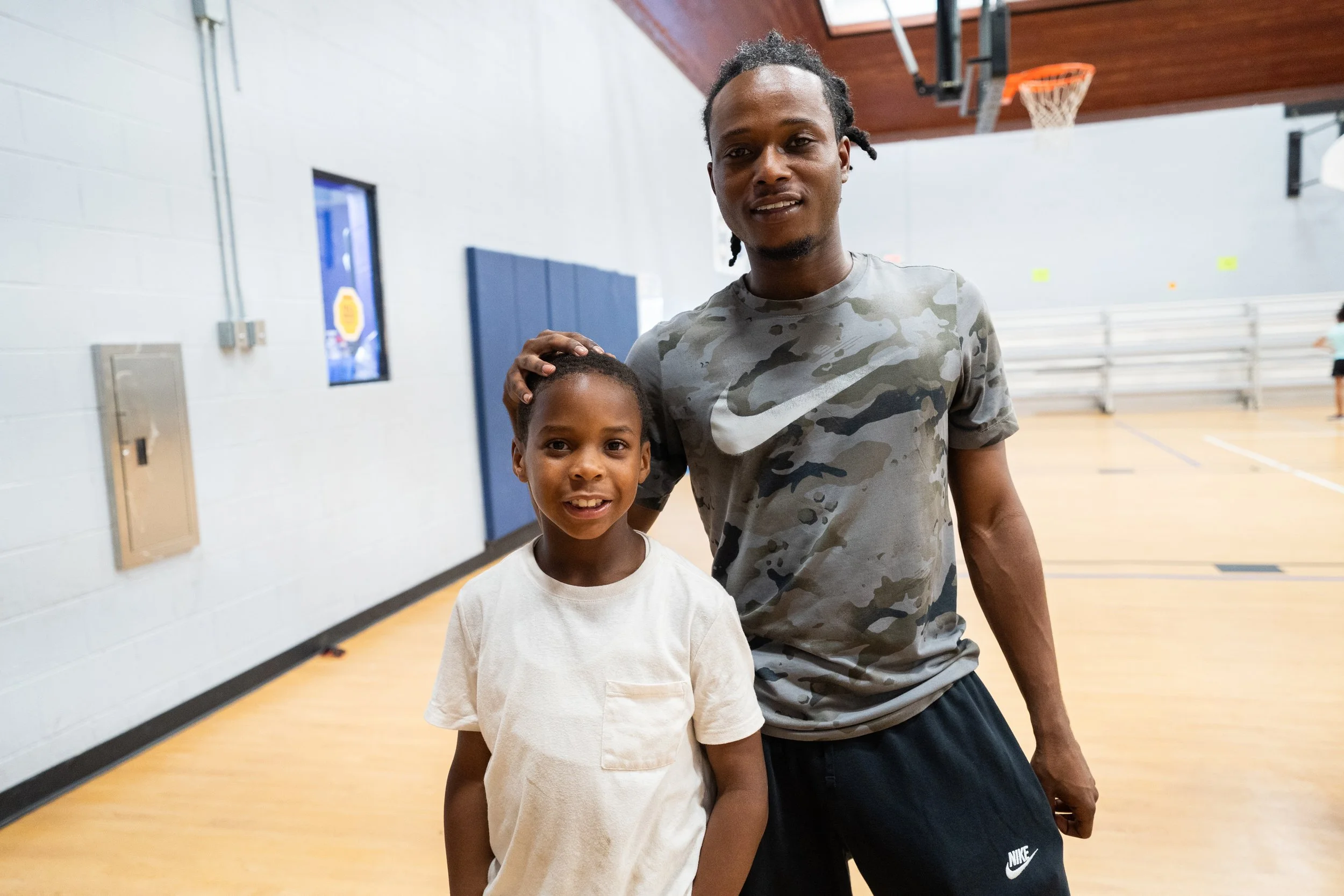 A young boy and a man standing in a basketball gym, with the man placing his hand on the boy's head and both smiling at the camera.