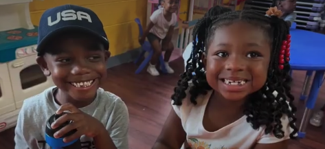 Two young children smiling and posing for the camera in a playroom filled with toys and furniture.