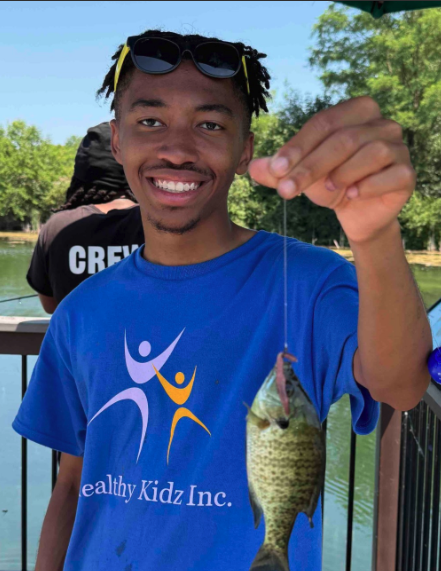 A young man holding a fishing line with a fish he caught, smiling, wearing sunglasses on his head and a blue t-shirt with a logo, outdoors near a lake with trees in the background.