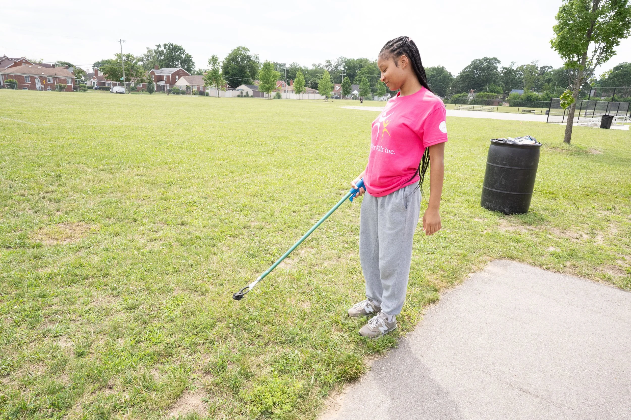 A young girl with braids, wearing a pink t-shirt and gray sweatpants, is using a grass trimmer at a park.