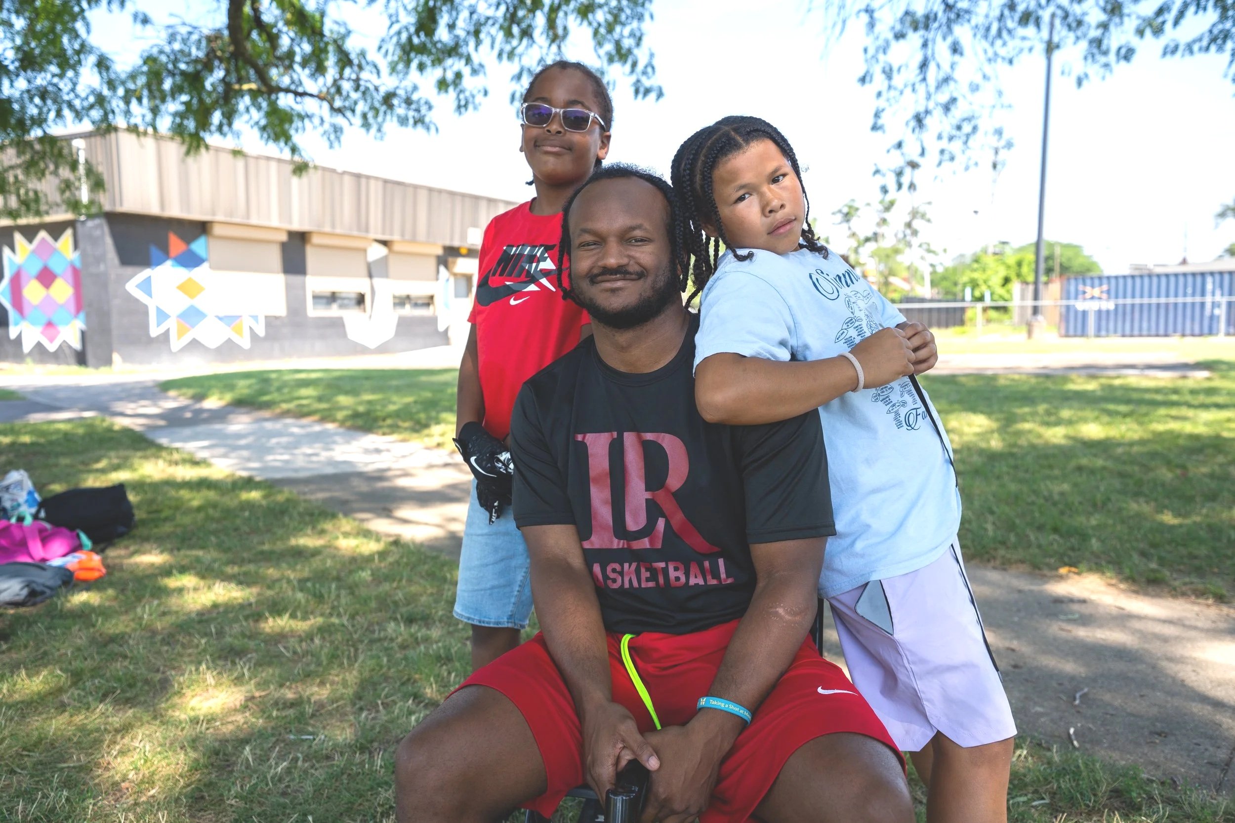 A man sitting outdoors on a chair with two young girls standing beside and behind him. The man has dreadlocks and is wearing a black T-shirt and red shorts. One girl is wearing sunglasses and a red shirt, standing behind him, and the other girl is wearing a light blue T-shirt and pink shorts, leaning on his shoulder. There are trees, a grassy area, and buildings in the background.