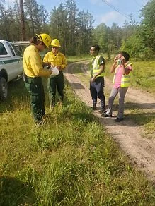 Three people in yellow uniforms and yellow helmets speaking to a woman with a pink shirt on a trail in a grassy area near trees.