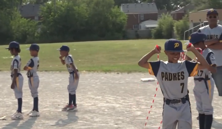 Young boys in baseball uniforms standing on a field, with one boy smiling and flexing his arms, others standing in line.