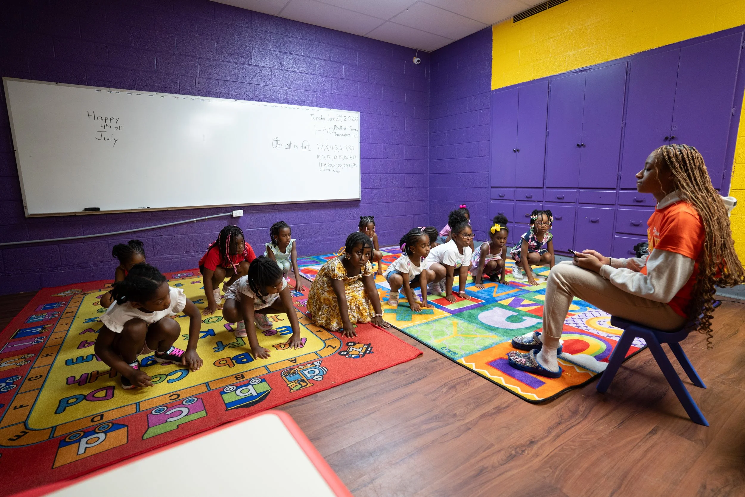 A teacher or instructor sitting on a chair in front of a group of young children in a classroom, instructing them as they perform a stretch or exercise on colorful alphabet rugs. The classroom has vibrant purple and yellow walls, a whiteboard with handwritten notes, and wooden flooring.