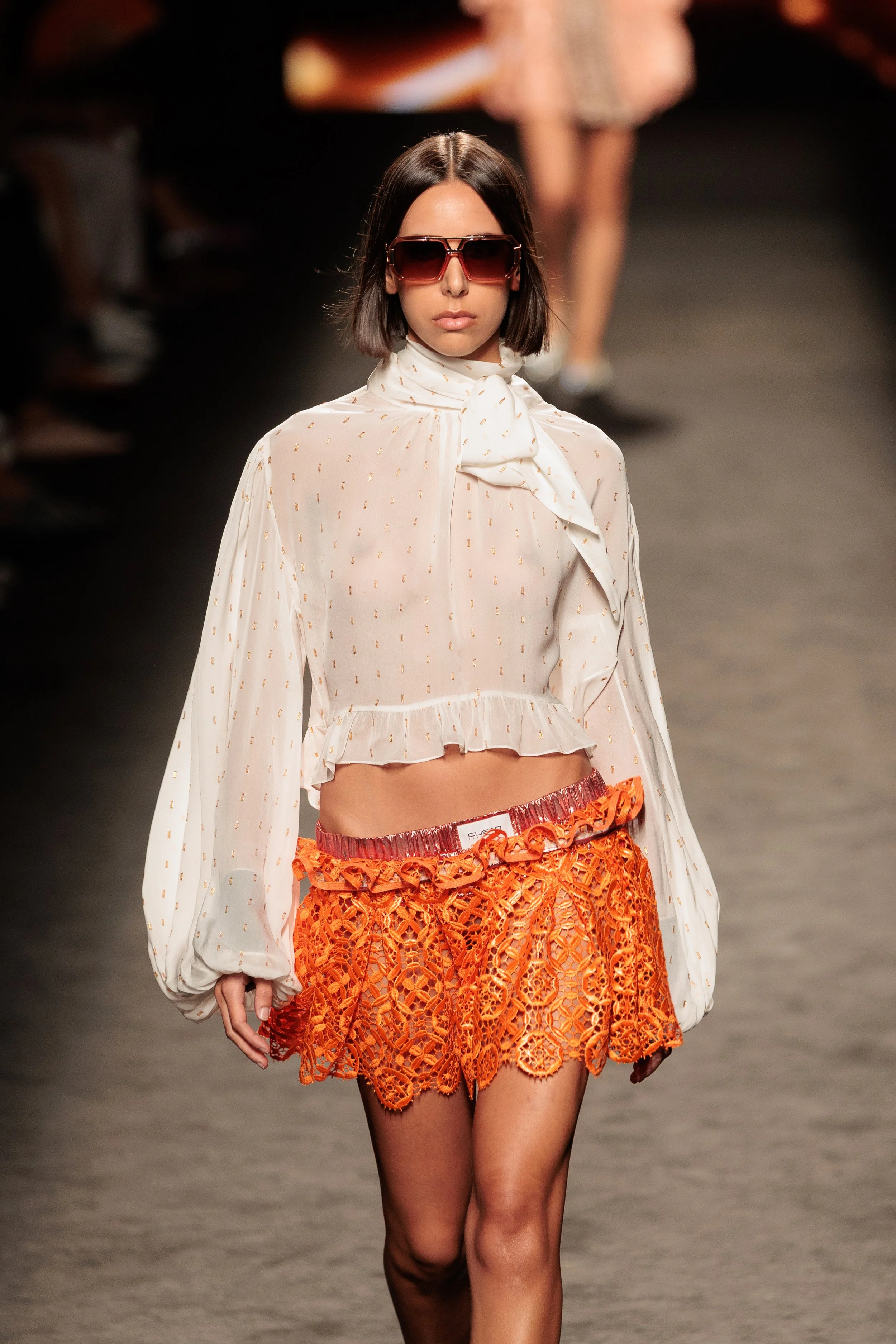 Modelo caminando en pasarela luciendo un conjunto de moda con blusa blanca de manga larga y hombros descubiertos, y falda naranja de encaje con detalles florales.