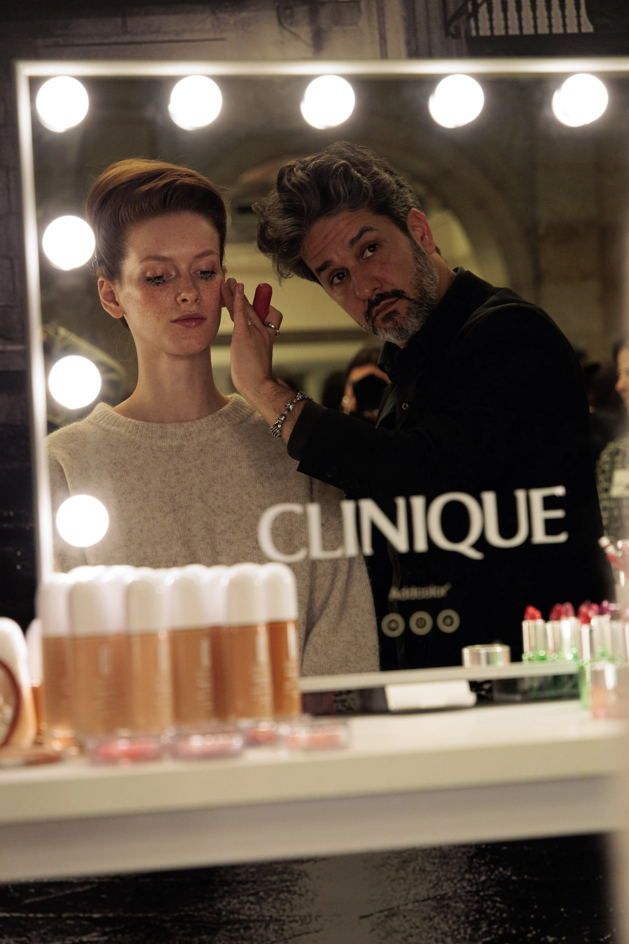 Un maquillador trabaja en una mujer con maquillaje y cabello arreglado en un espejo de tocador que lleva la marca "Clinique" en un entorno de sala de preparación o estudio.