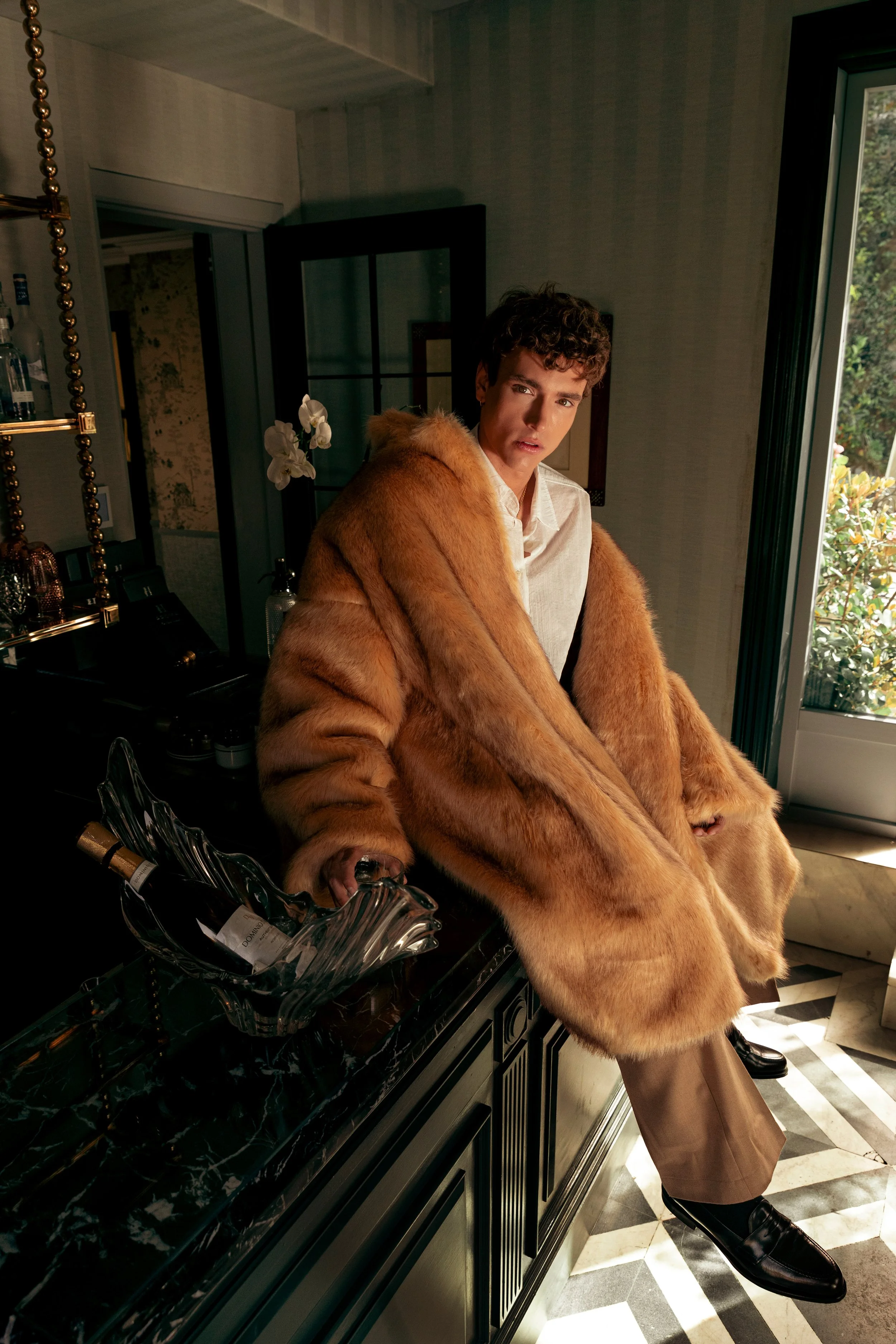 Joven con abrigo de piel dorada, de pie junto a una barra de bar en una sala con luz natural y plantas en el fondo.