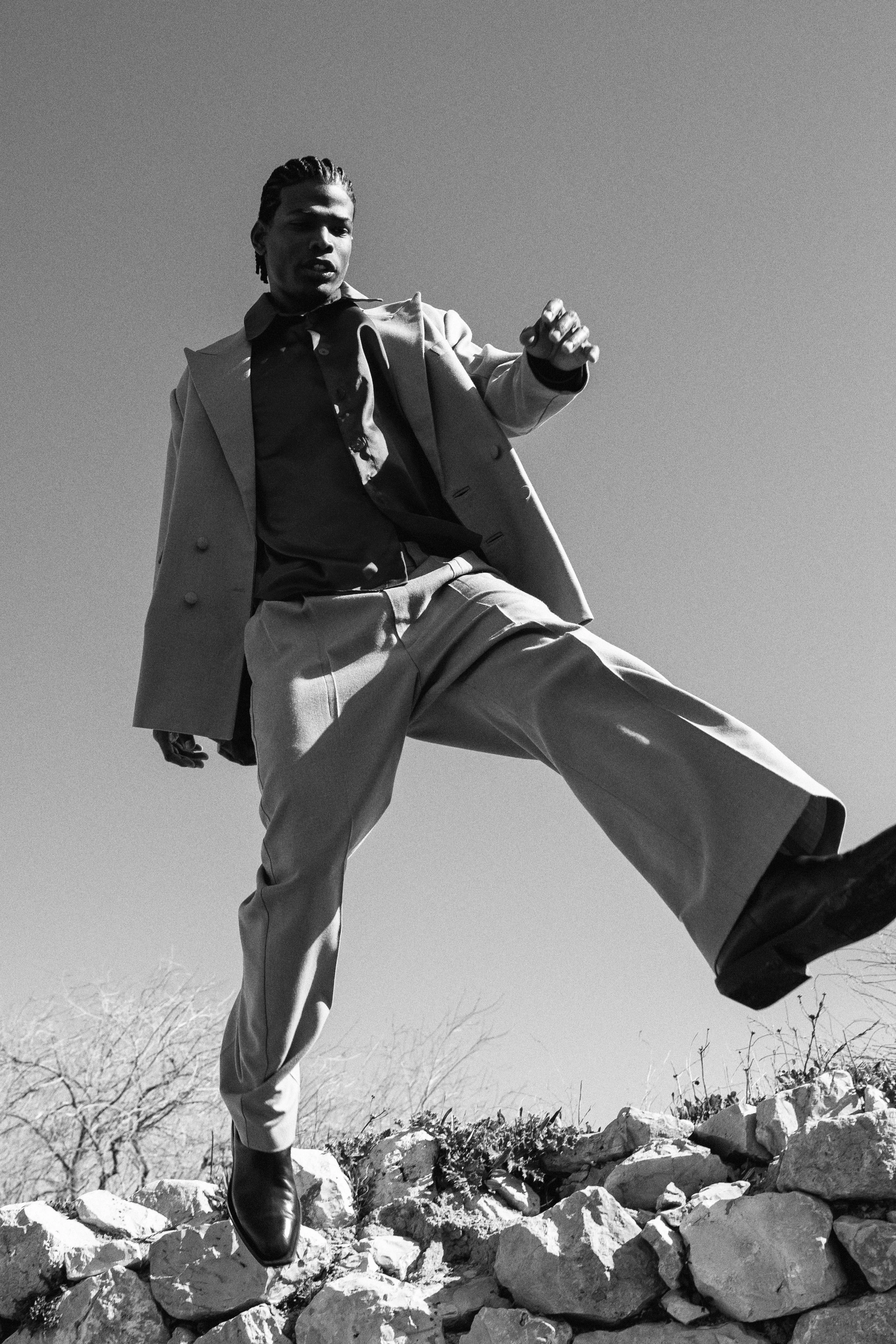 Hombre vestido con ropa formal saltando sobre piedras en un paisaje exterior en blanco y negro.