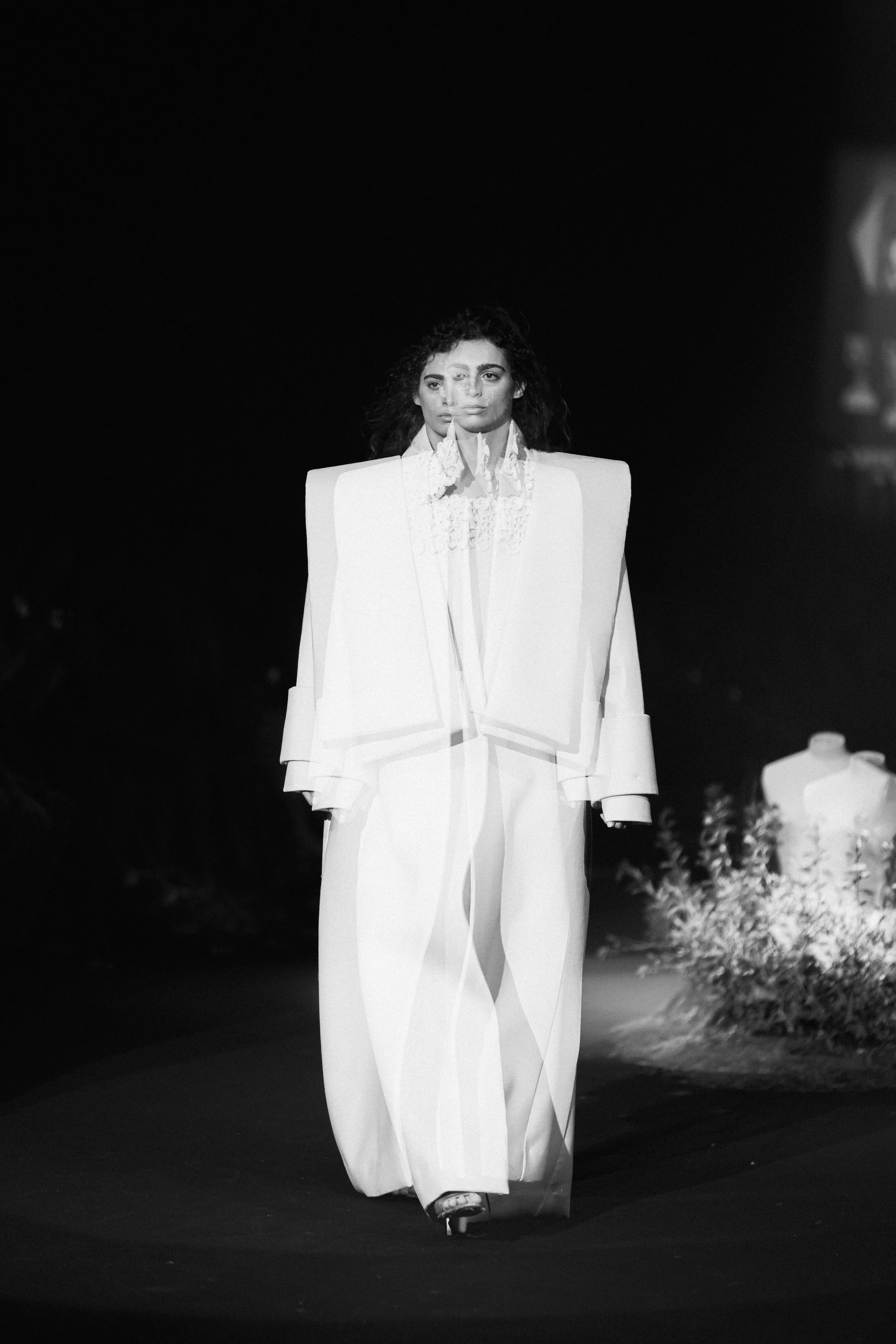 Modelo caminando en pasarela durante un desfile de moda, vestida con un traje blanco y fondo oscuro.