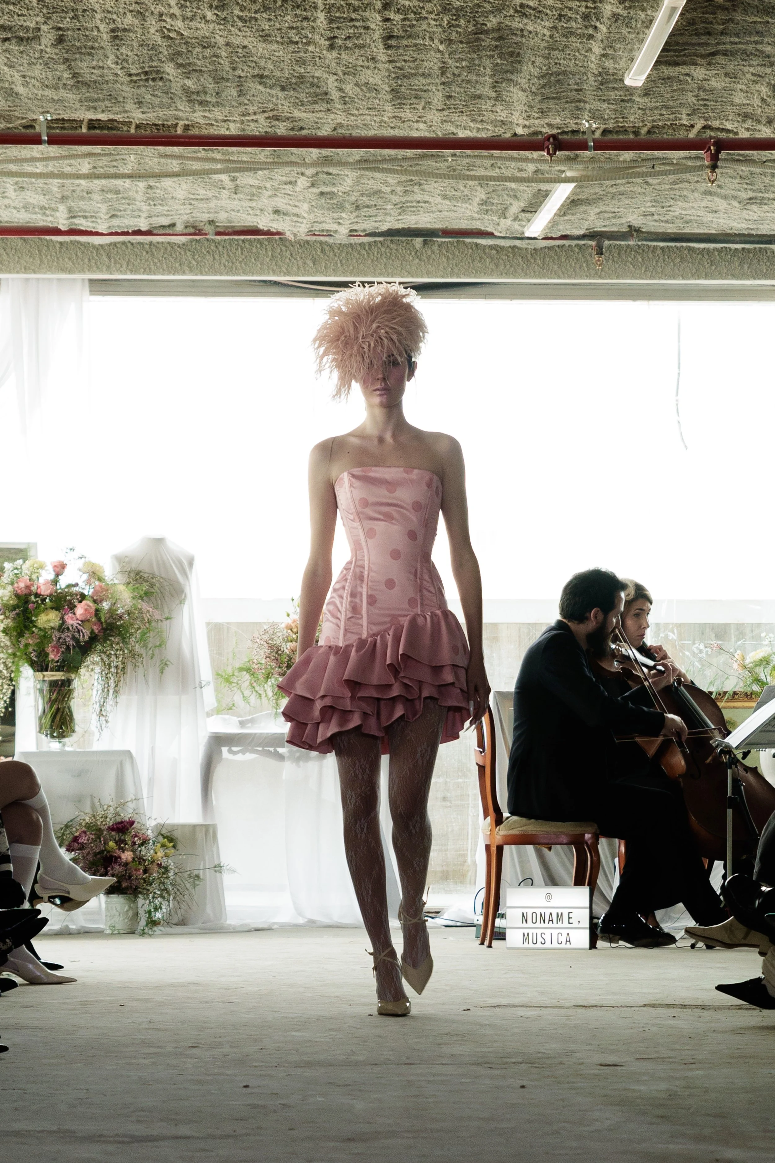 Modelo caminando en pasarela con vestido rosa, peinado con peluca peluda, en un escenario con músicos y arreglos florales.