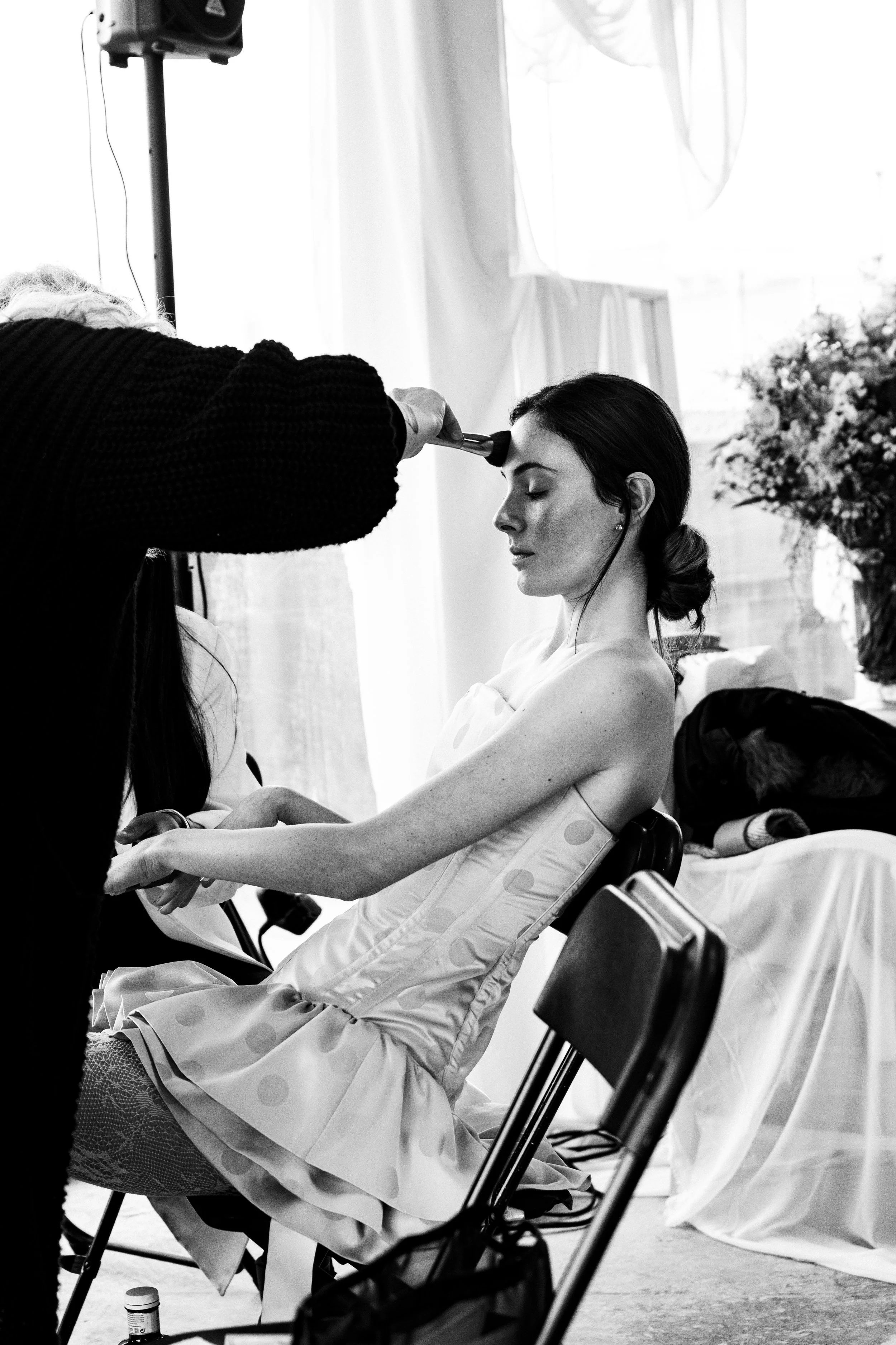 Una mujer en un vestido sentado con los ojos cerrados, recibiendo maquillaje o arreglo en un evento.