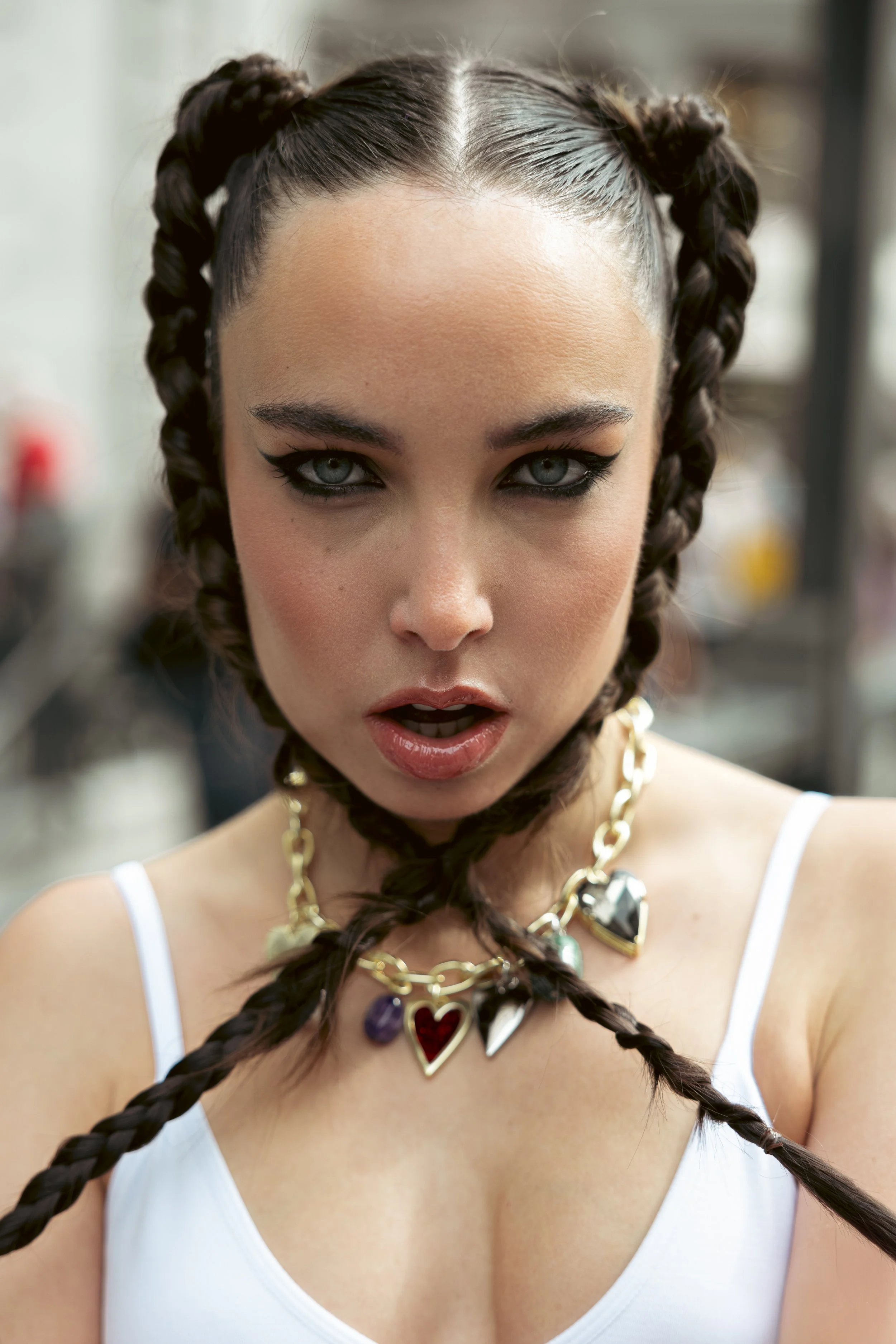 Retrato cercano de una mujer con cabello rizado en dos coletas, maquillaje llamativo, collar con corazones y objetos colgantes, usando camiseta blanca.