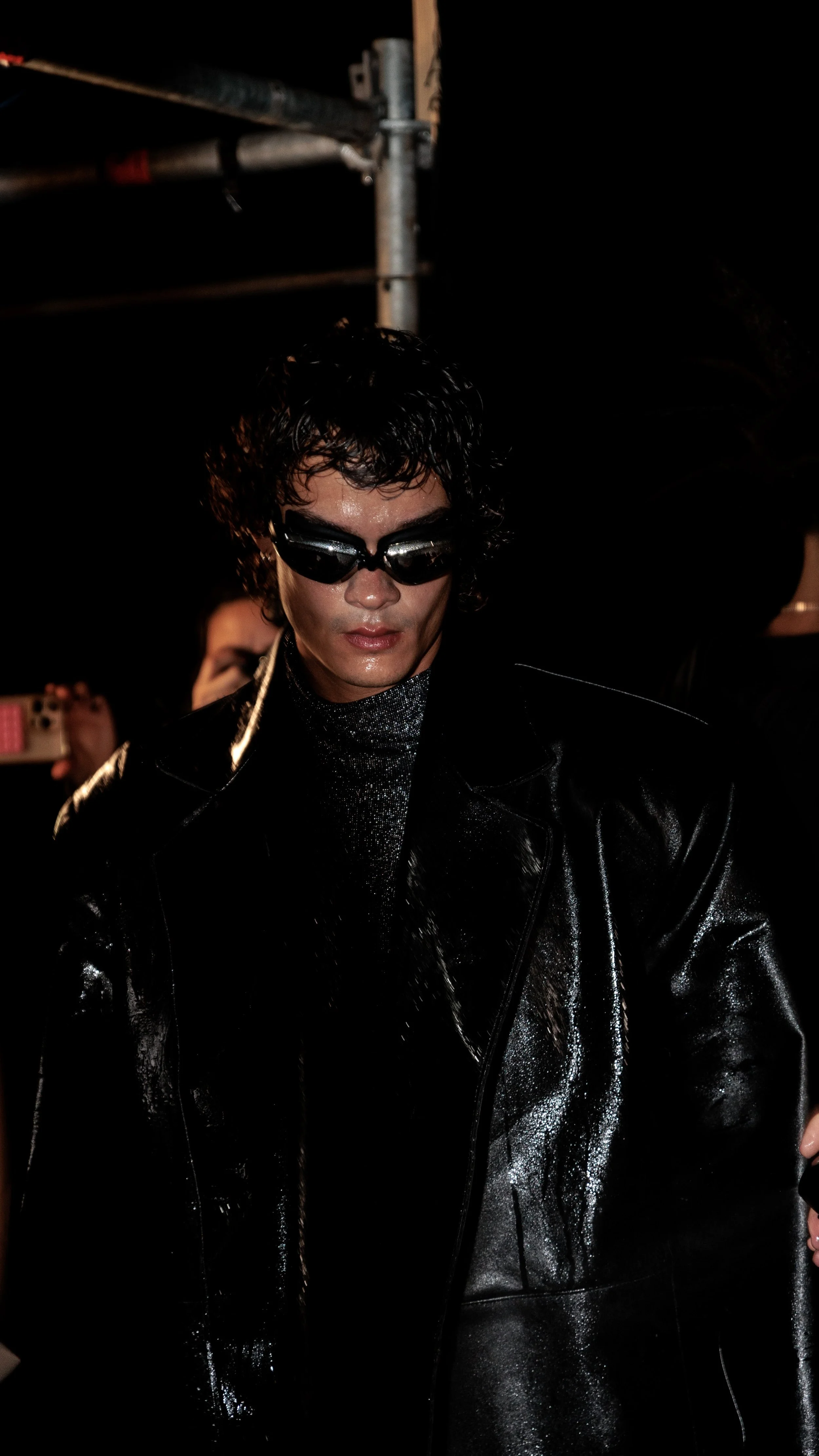 Hombre con pelo rizado, gafas oscuras, chaqueta de cuero negra y ropa brillante, en un entorno nocturno con iluminación tenue.