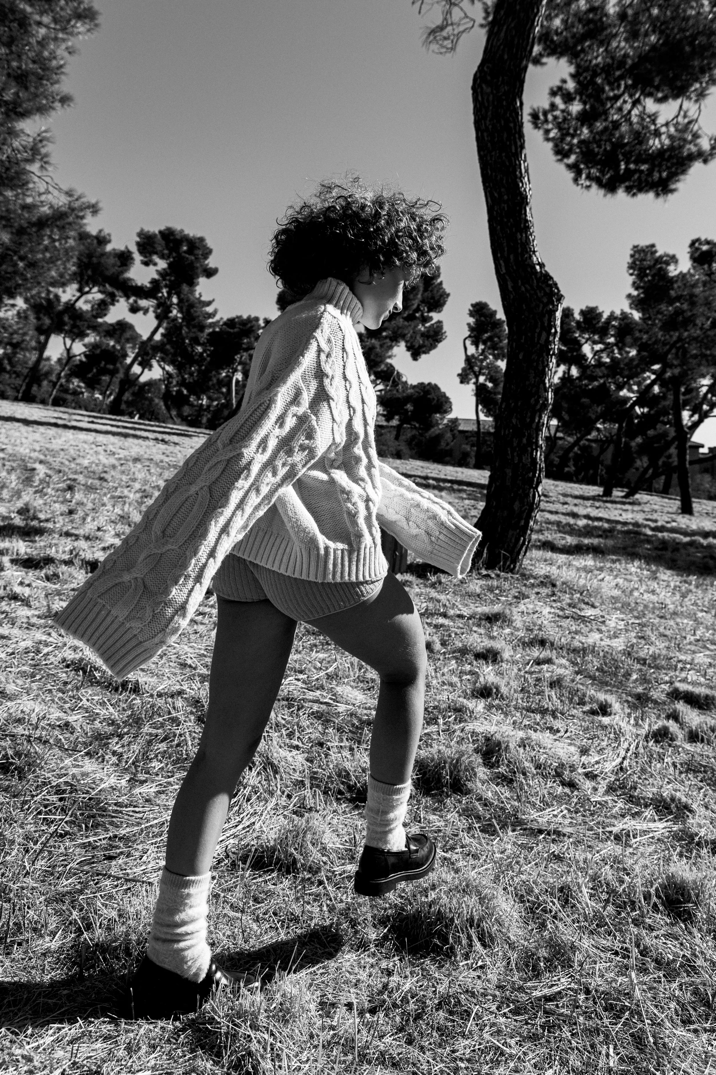 Una niña de cabello rizado caminando por un campo con árboles, vista en blanco y negro.