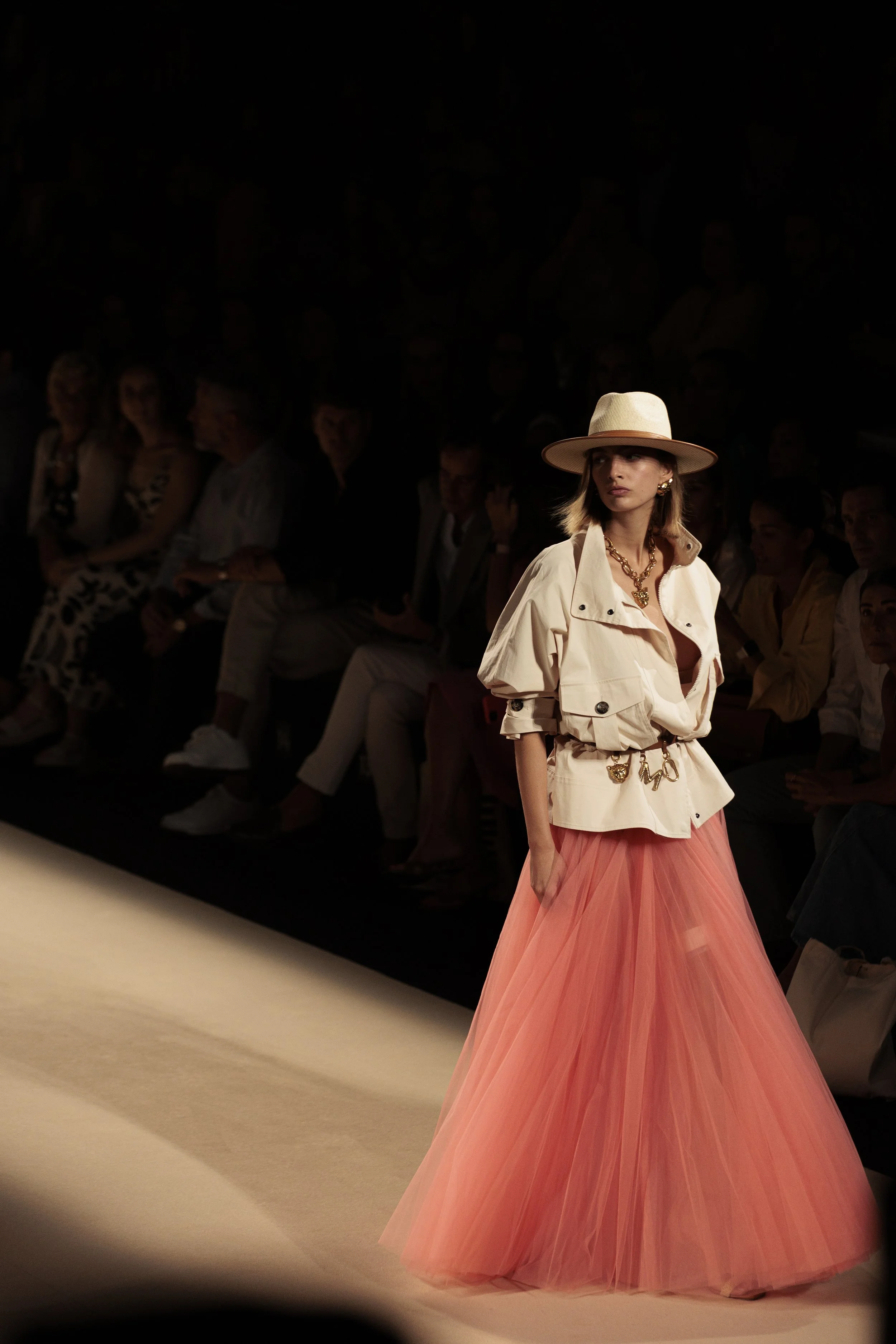 Modelo desfila con sombrero, chaqueta beige y falda larga de tule rosada en pasarela de moda.