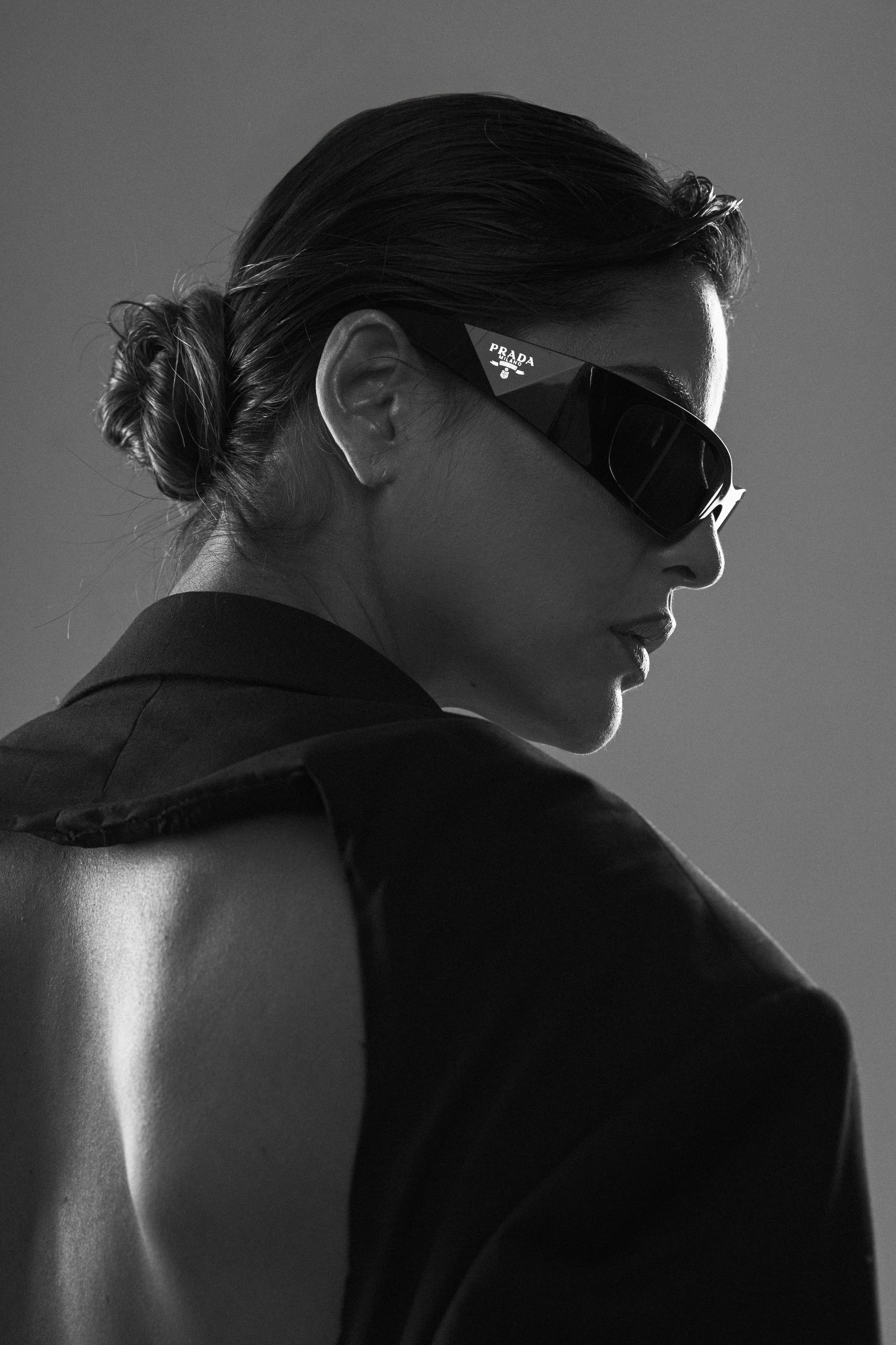 Retrato en blanco y negro de una mujer con peinado en moño, gafas de sol Prada, vestimenta oscura y fondo simple.