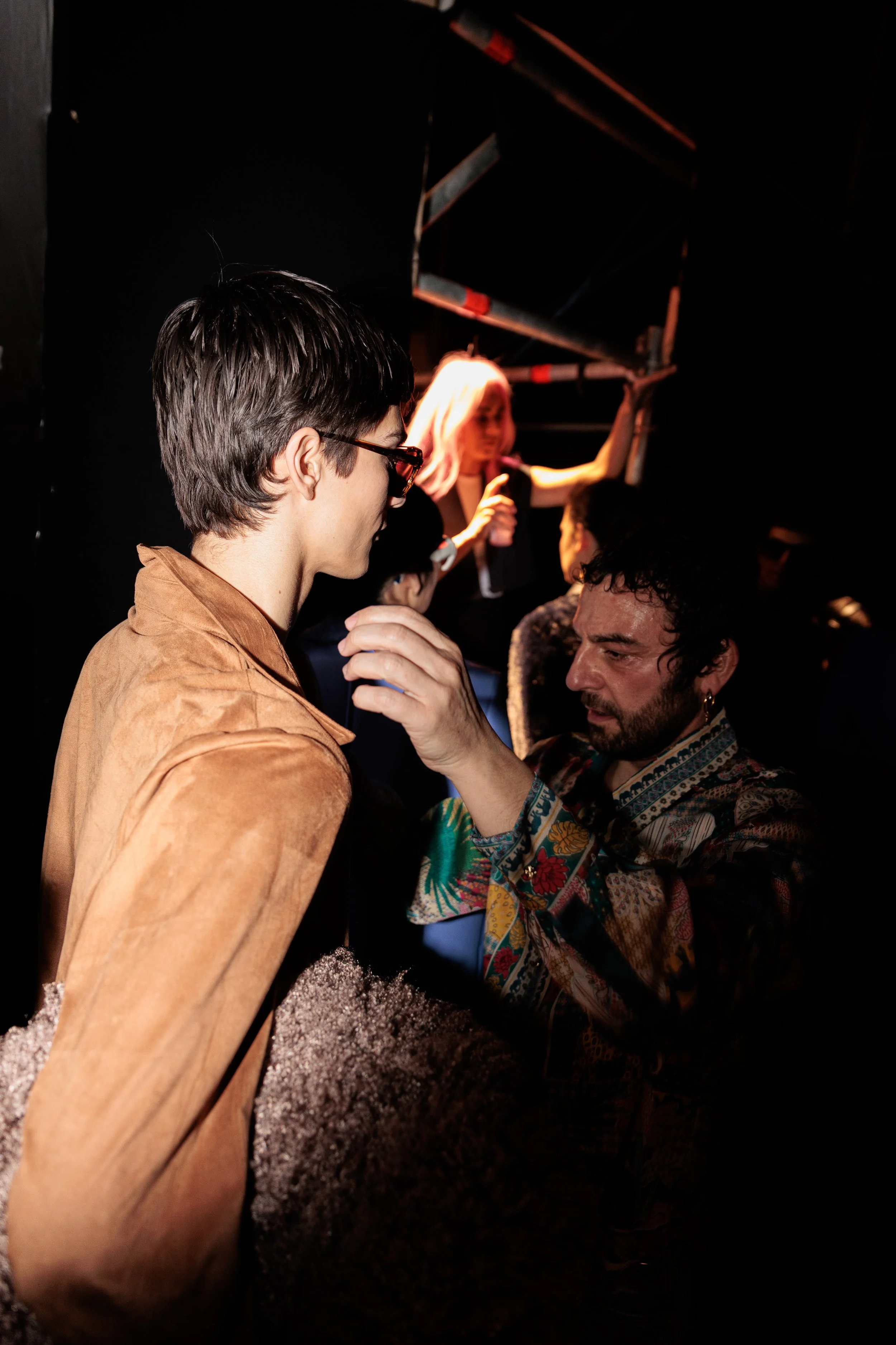 Dos personas en un espacio oscuro, aparentemente en un evento o concierto. Una de ellas, con gafas y cabello corto, mira mientras otra persona ajusta su collar o oreja. En el fondo, hay una mujer rubia con cabello suelto sosteniéndose de una barra o 