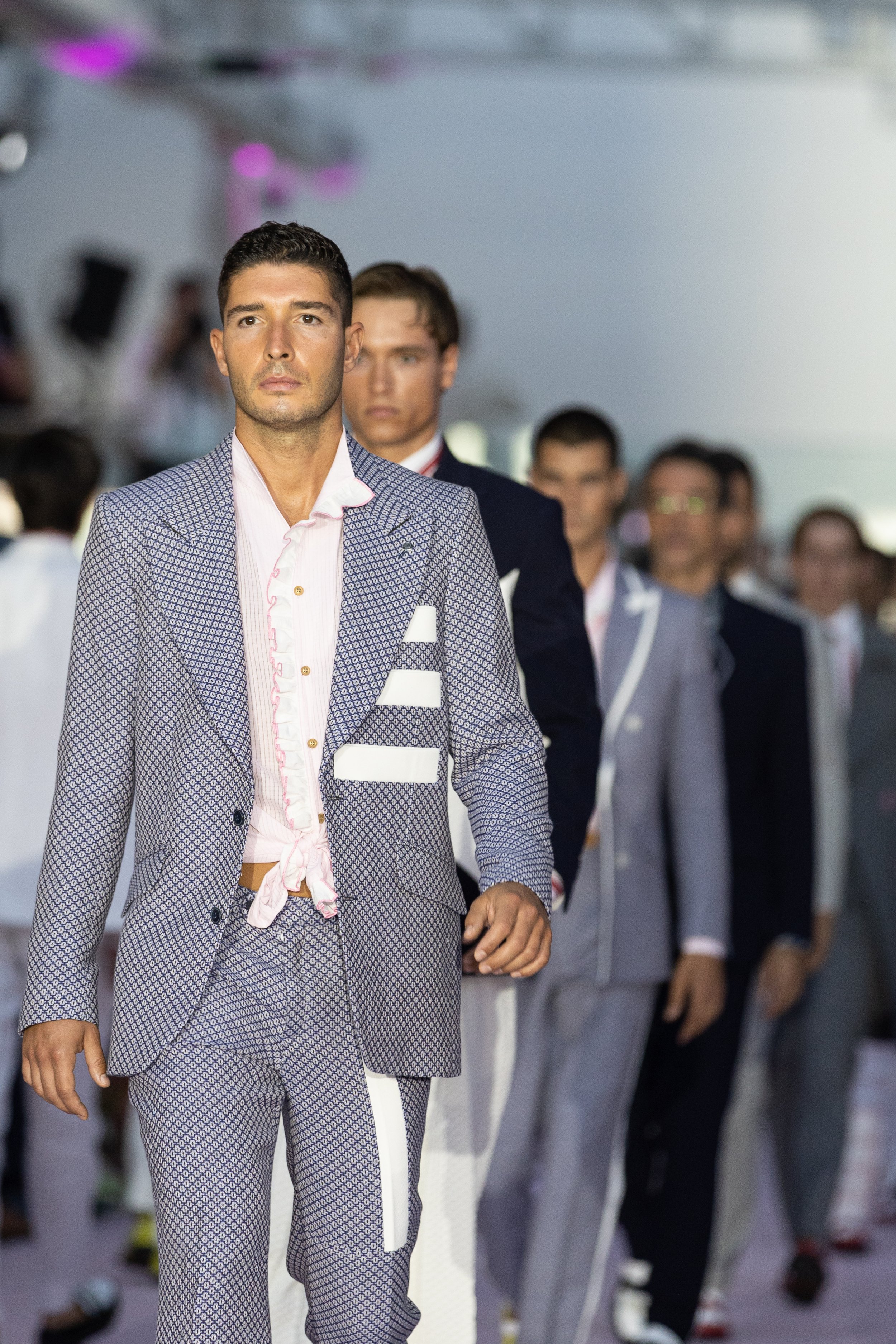 Hombres desfilando en pasarela de moda en un evento con fondo difuso y luces.