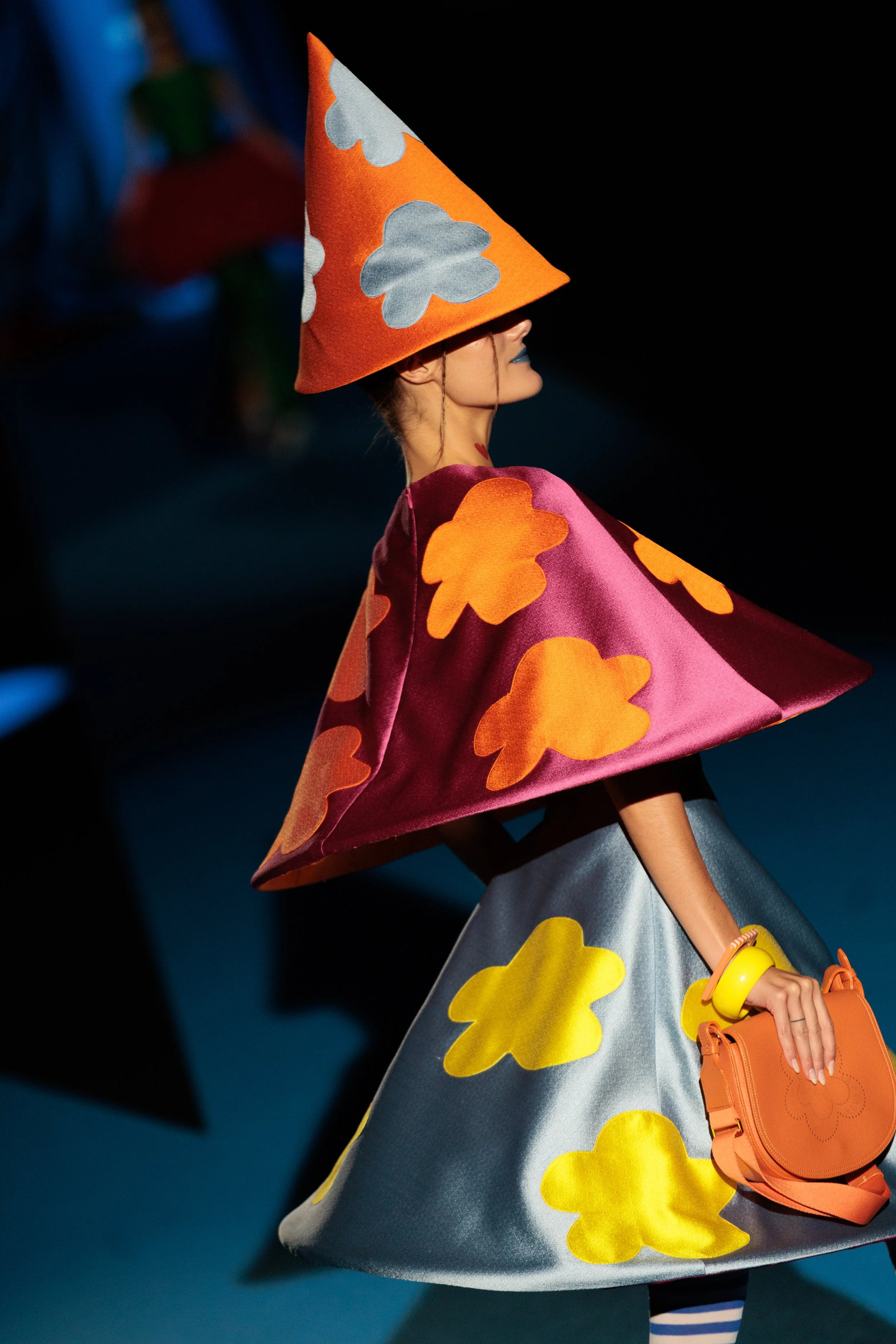 Modelo caminando en una pasarela con vestuario colorido y accesorios llamativos durante un desfile de moda.