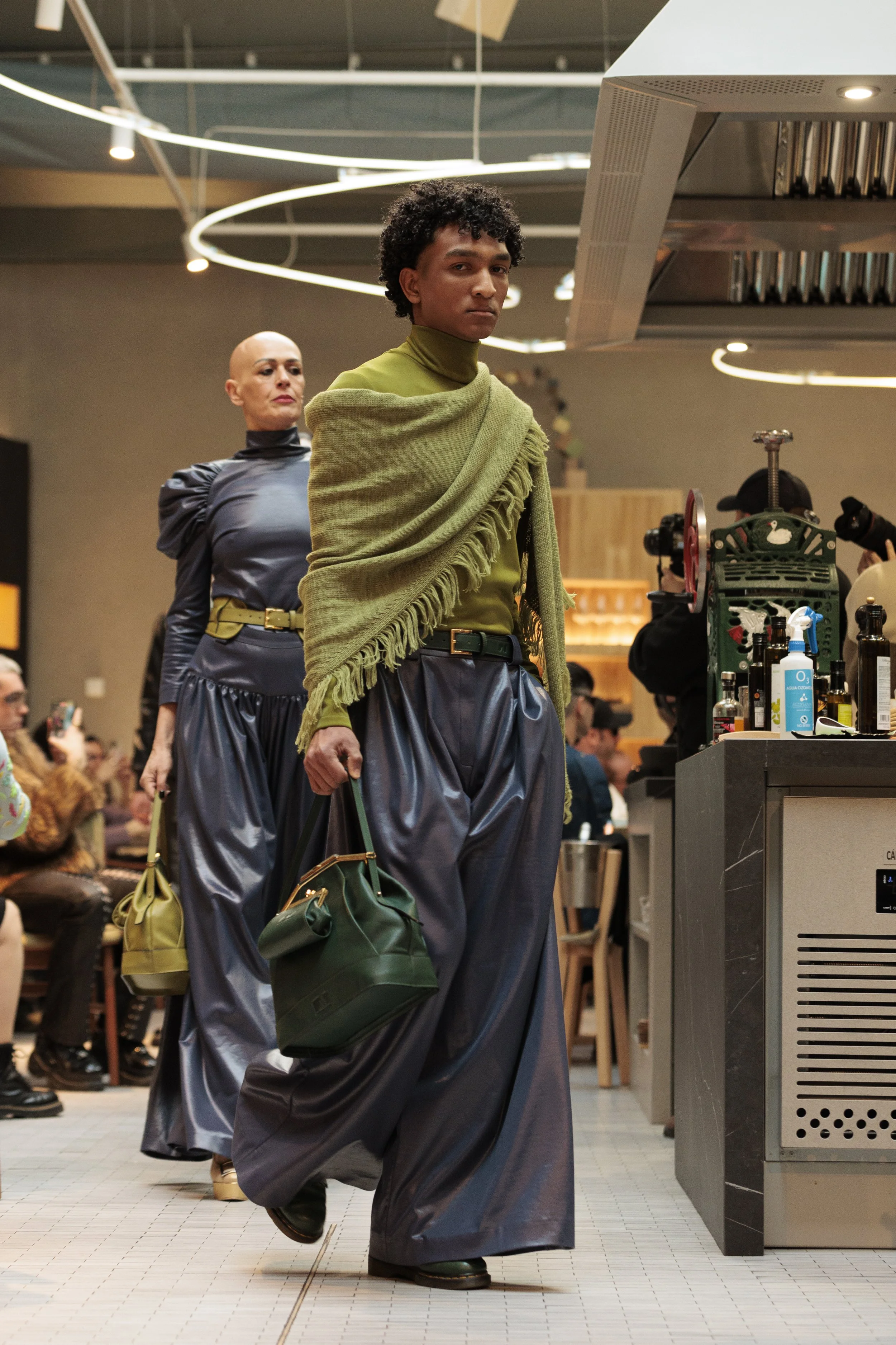 Dos personas con ropa moderna caminando en un lugar público, una lleva una prenda de color verde y una bolsa verde, la otra lleva un vestido oscuro y una bolsa amarilla, en un ambiente concurrido con gente y decoración moderna.