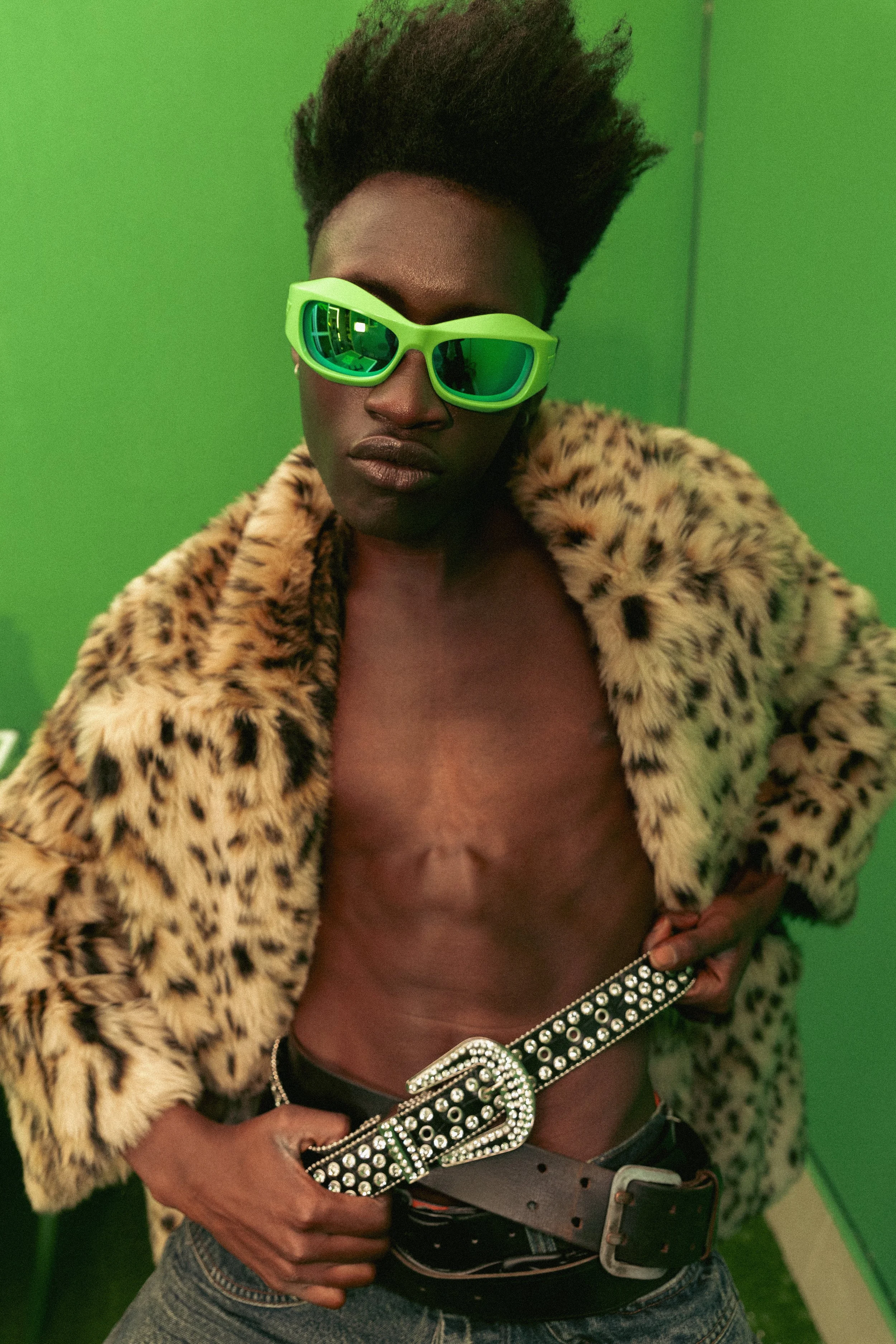 Persona con gafas verdes, chaqueta de peluche de leopardo, cinturón con adornos de perlas y sin camisa, en un fondo verde.