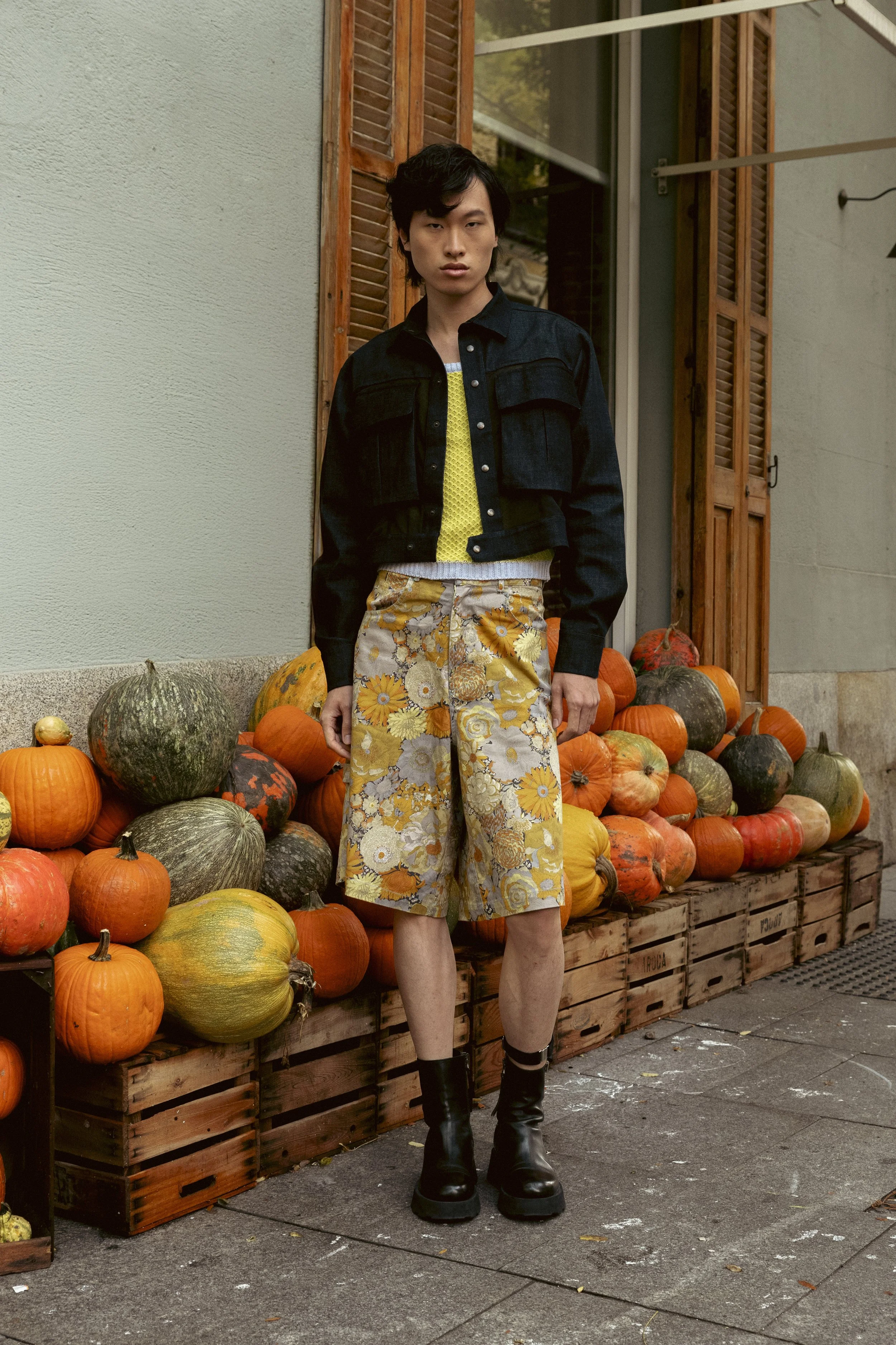 Persona joven con cabello negro, vestida con chaqueta de mezclilla negra, falda corta estampada con flores en tonos amarillos y naranjas, botas negras, de pie frente a una exhibición de calabazas de diferentes tamaños y colores en un puesto callejero