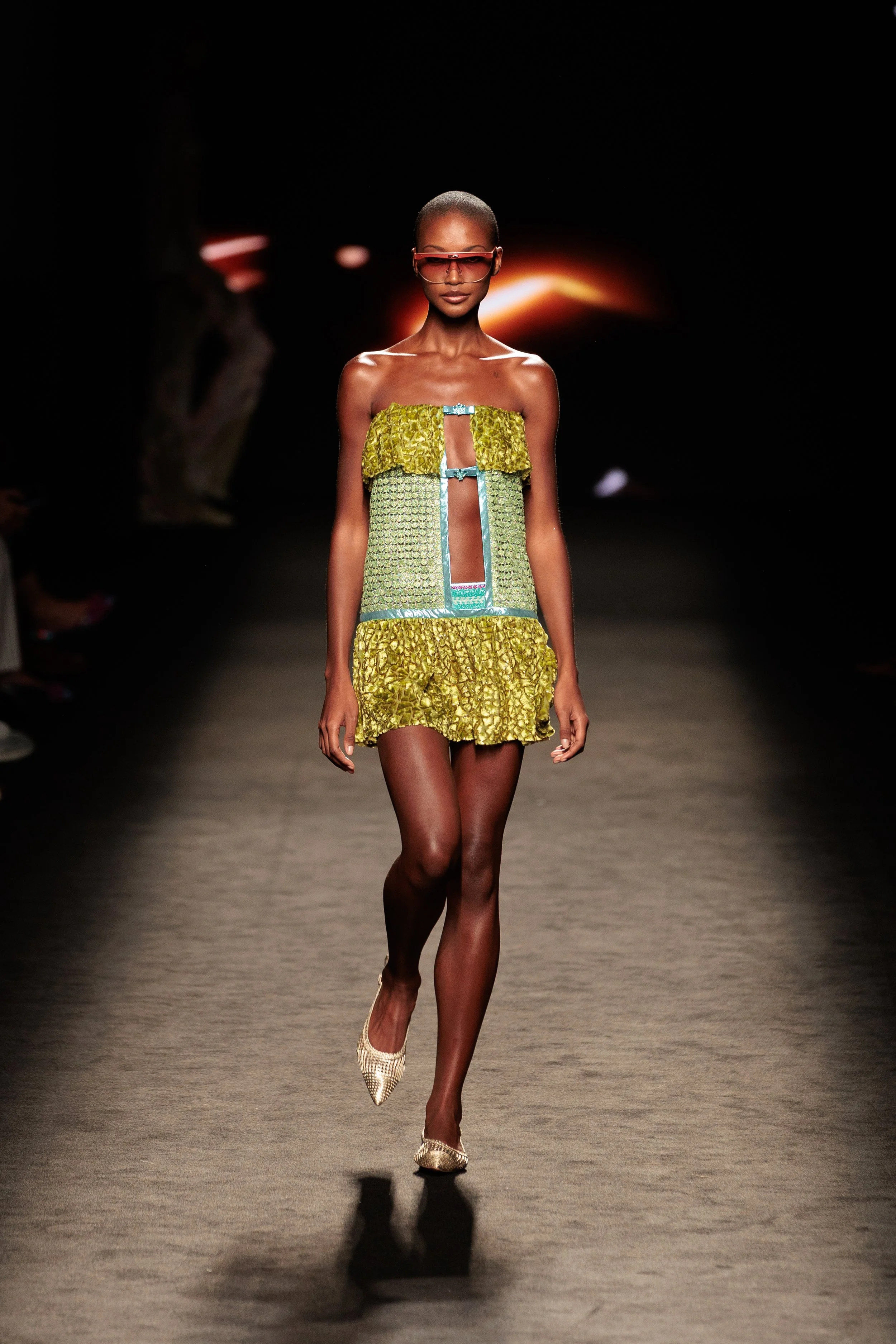 Modelo femenina caminando en pasarela con vestido color verde y accesorios de gafas de sol rosadas en fondo oscuro con luz de puesta de sol.