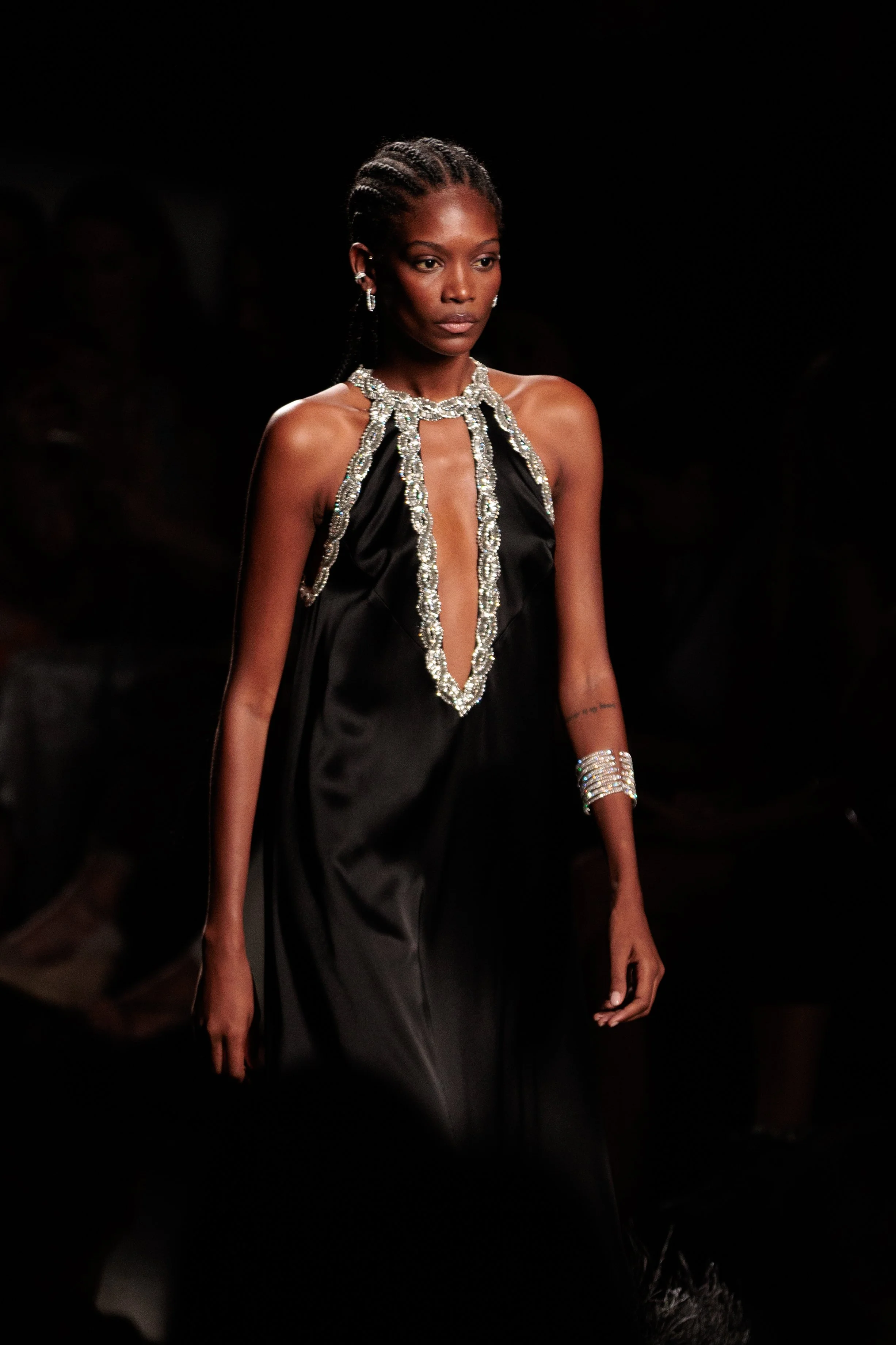 Mujer caminando en pasarela con vestido largo negro, adornado con piedras y pedrería, en un desfile de moda