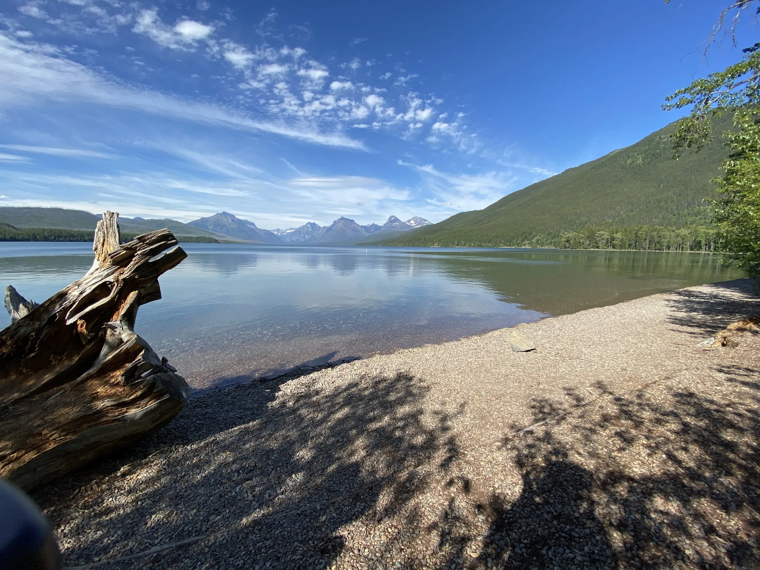 A peaceful lake with clear water reflecting blue skies and clouds, surrounded by green mountains, with a sandy beach and a large piece of driftwood in the foreground.