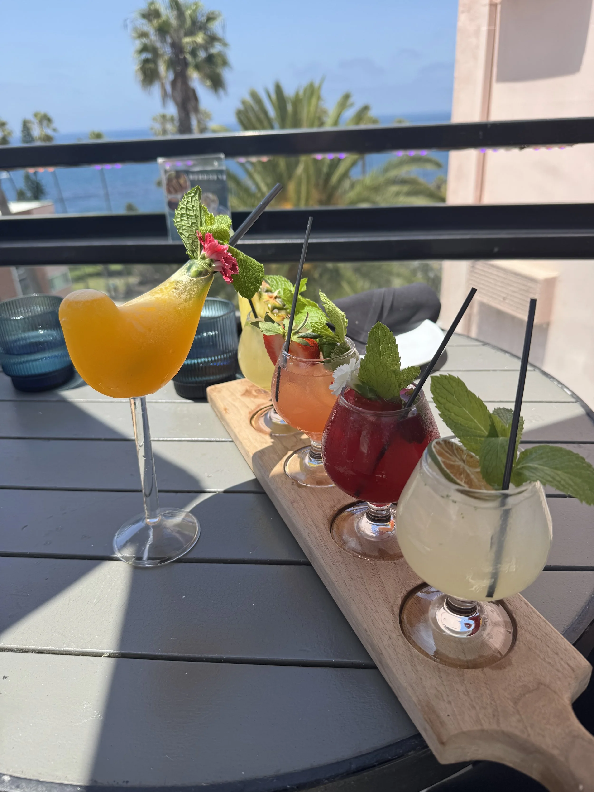 A line-up of colorful cocktails with garnishes on a wooden serving board, set on a table with a view of palm trees, a building, and the ocean in the background.