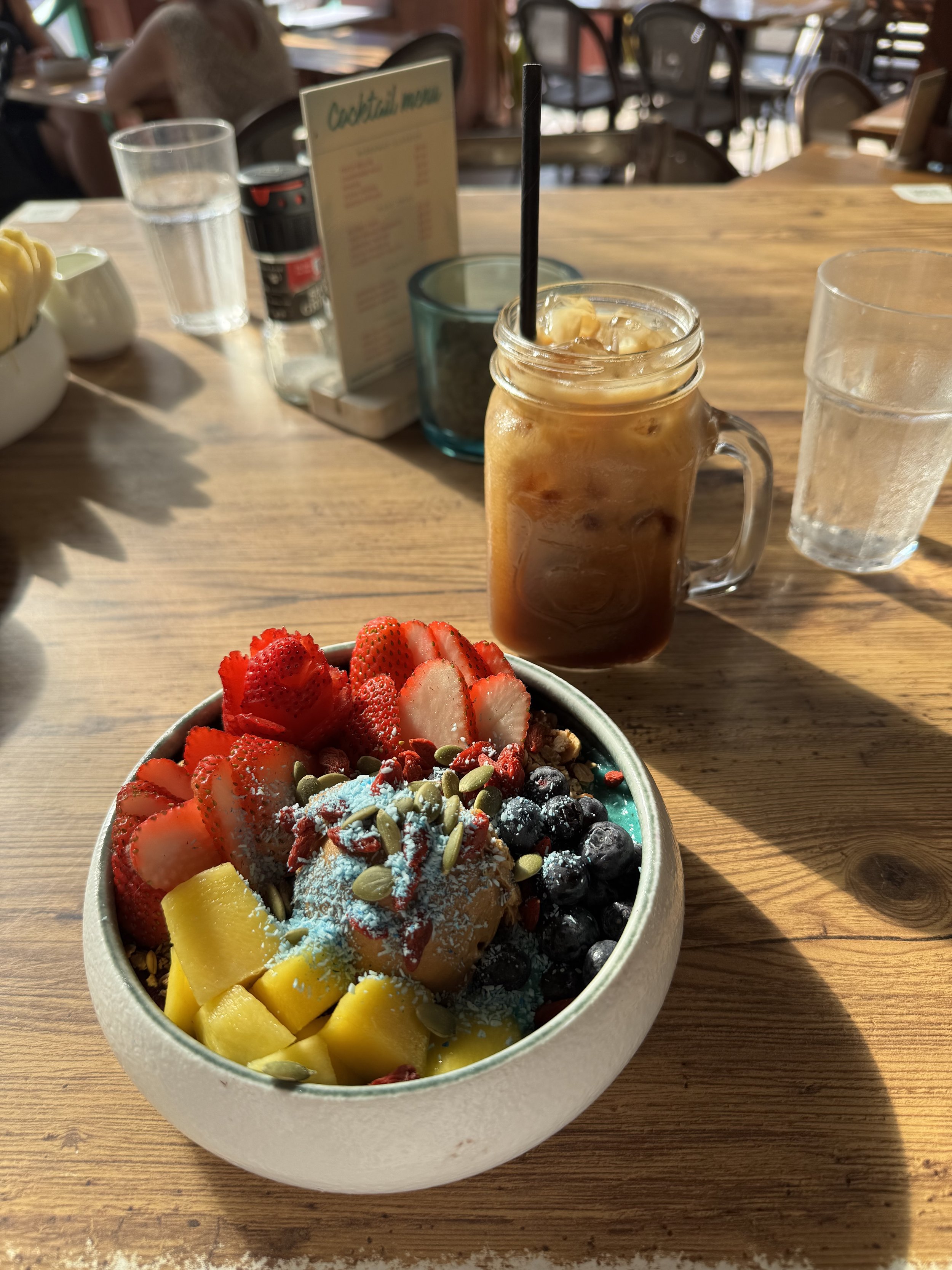 A bowl of acai fruit, topped with strawberries, blueberries, mango, pumpkin seeds, and a scoop of ice cream or frozen yogurt, accompanied by an iced coffee in a mason jar and two glasses of water on a wooden table in a restaurant.