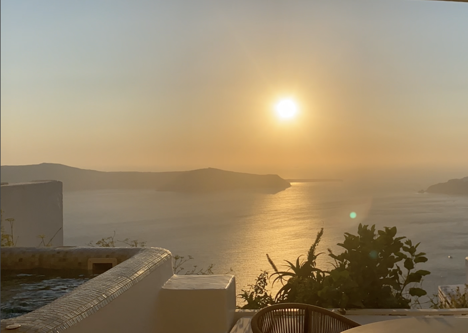 Sunset over a calm sea with distant islands, seen from a patio with an outdoor hot tub, plants, and a chair in the foreground.