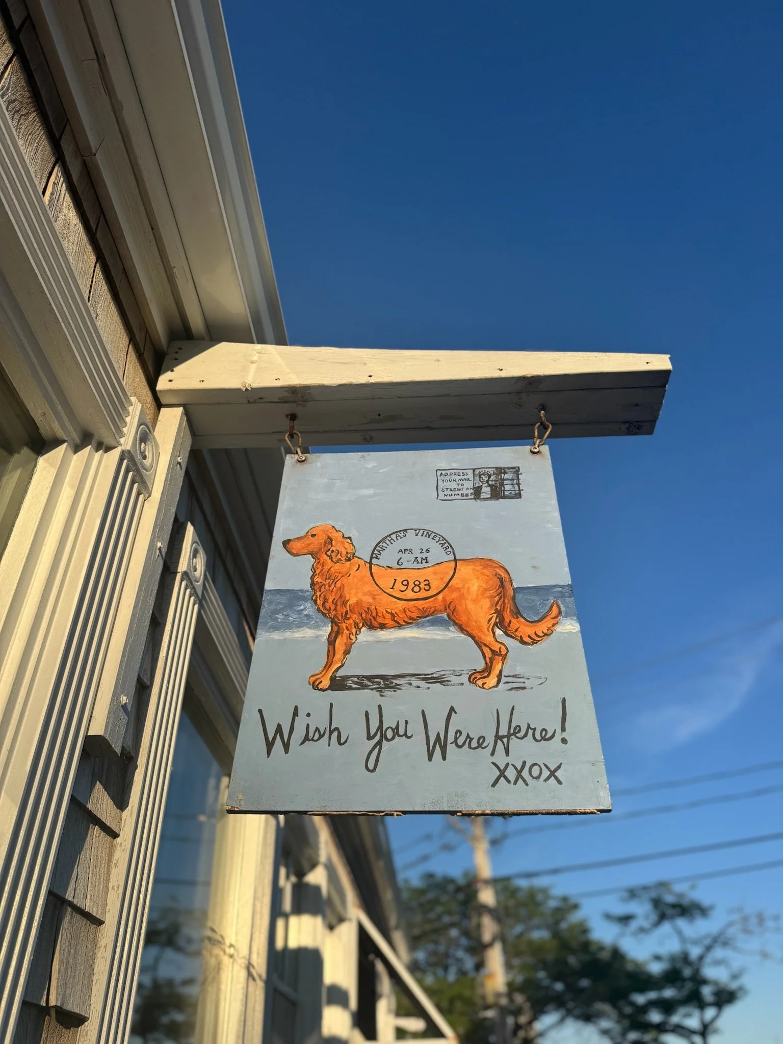 A decorative sign hanging outside a building features an illustration of an orange dog on a beach background. The sign includes the text "Wish You Were Here! XXOX" and has a vintage stamp with the date 1983 and the location Northas Vineyard. The sign